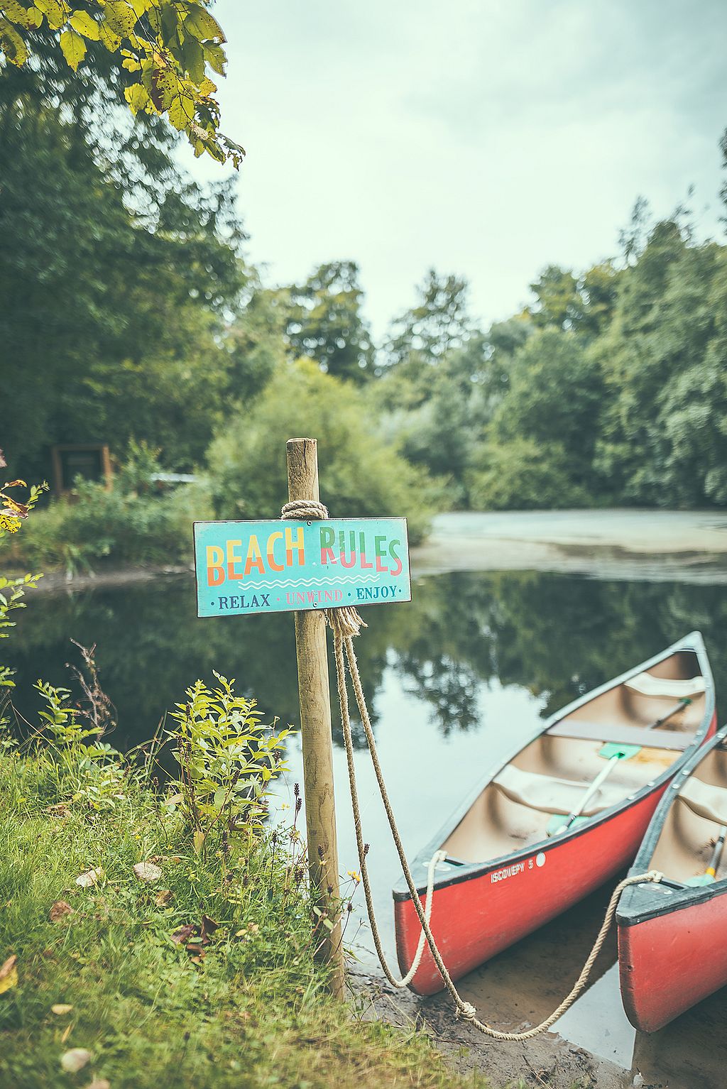 A colourful ‘Beach Rules’ sign stands beside two red canoes pulled up at the edge of a calm wooded lake.