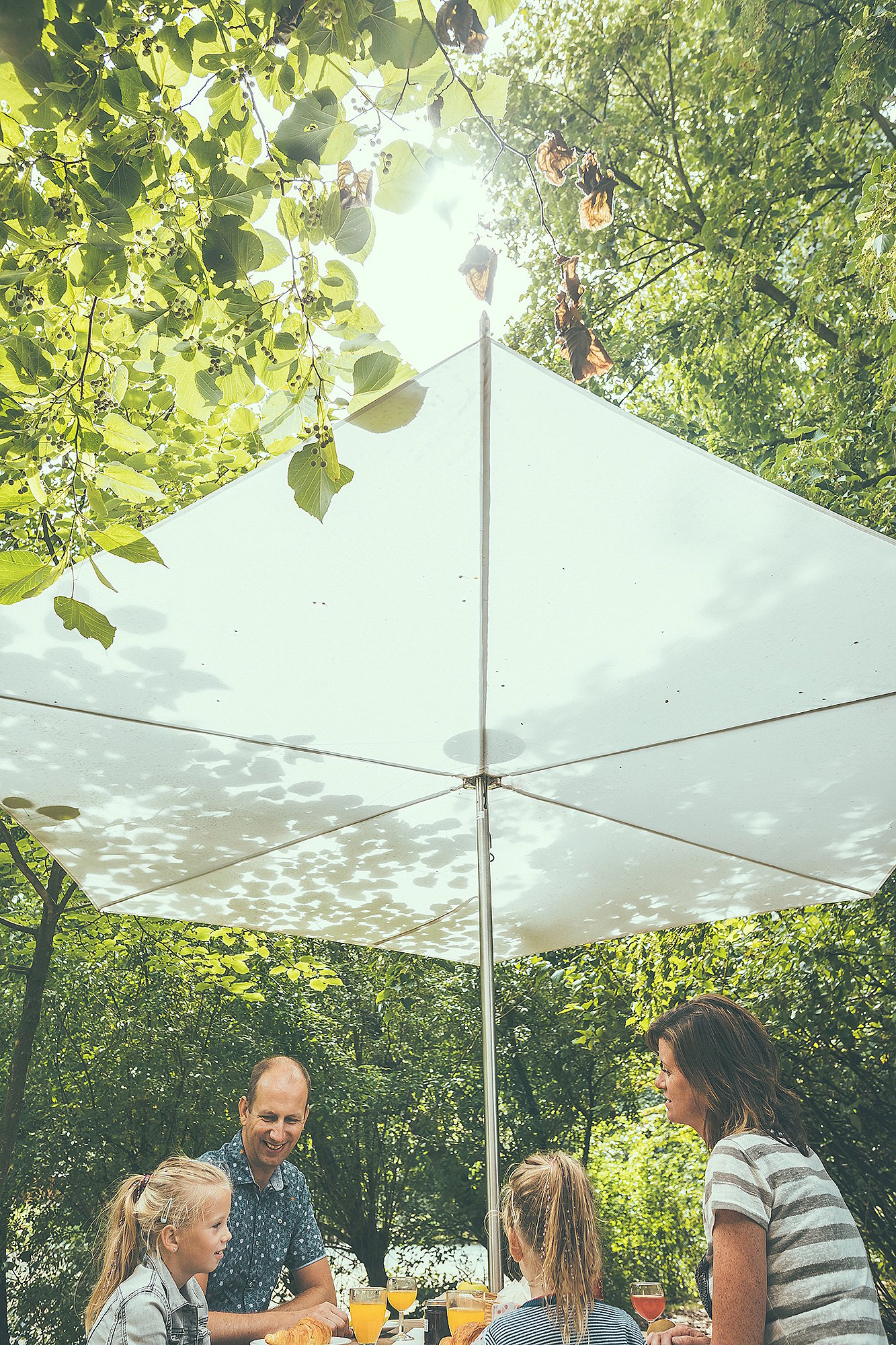 Low-angle view of a white Inumbrina parasol shading a family gathered around an Anker picnic table in a wooded clearing.