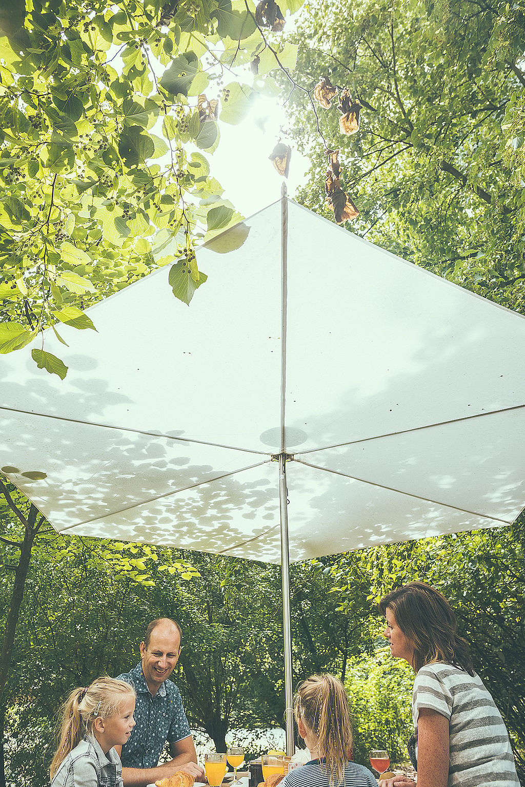 Low-angle view of a white Inumbrina parasol shading a family gathered around an Anker picnic table in a wooded clearing.