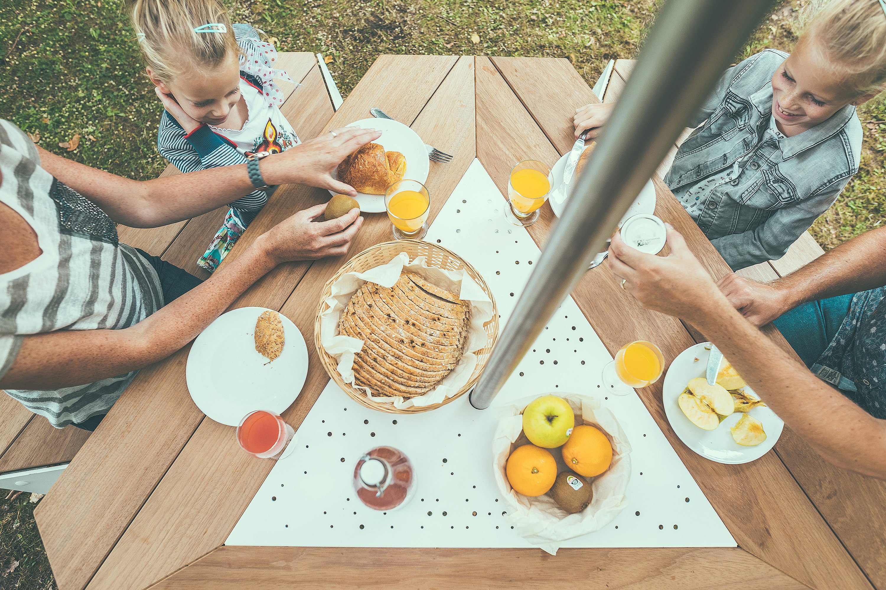 Overhead view of breakfast plates, fruit and pastries on the wooden tabletop of an Anker picnic table beneath the central pole of an Inumbrina parasol.
