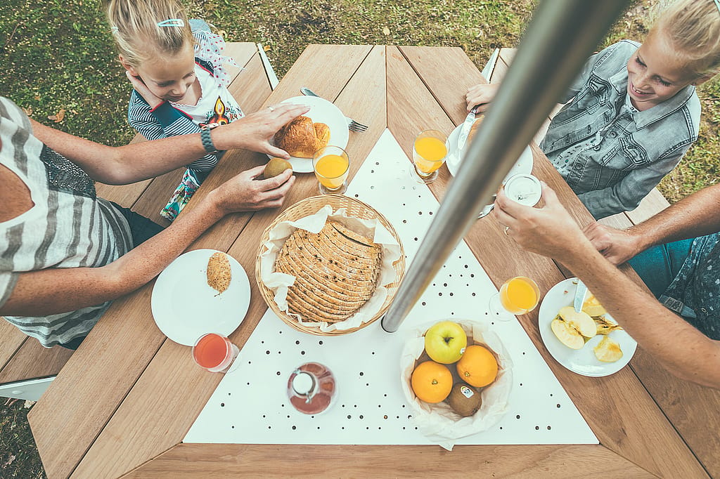 Overhead view of breakfast plates, fruit and pastries on the wooden tabletop of an Anker picnic table beneath the central pole of an Inumbrina parasol.