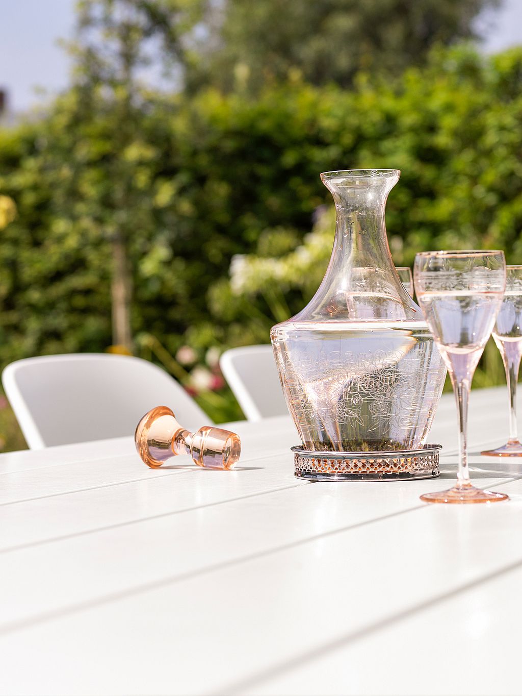 Nahaufnahme einer Glaswasserkaraffe und hellen Stielgläsern auf einer weißen Tischplatte, wobei ein Glas auf der Seite liegt und dahinter ein üppig grüner Garten sanft verschwimmt