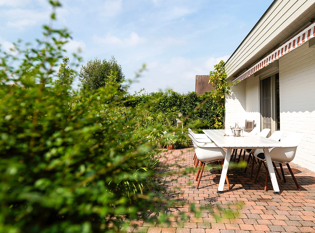 Weißer Esstisch im Yachthafen im Freien und passende Kapitänsstühle auf einer Backsteinterrasse neben einem weißen Backsteinhaus, gesehen durch weiches Gartenlaub.
