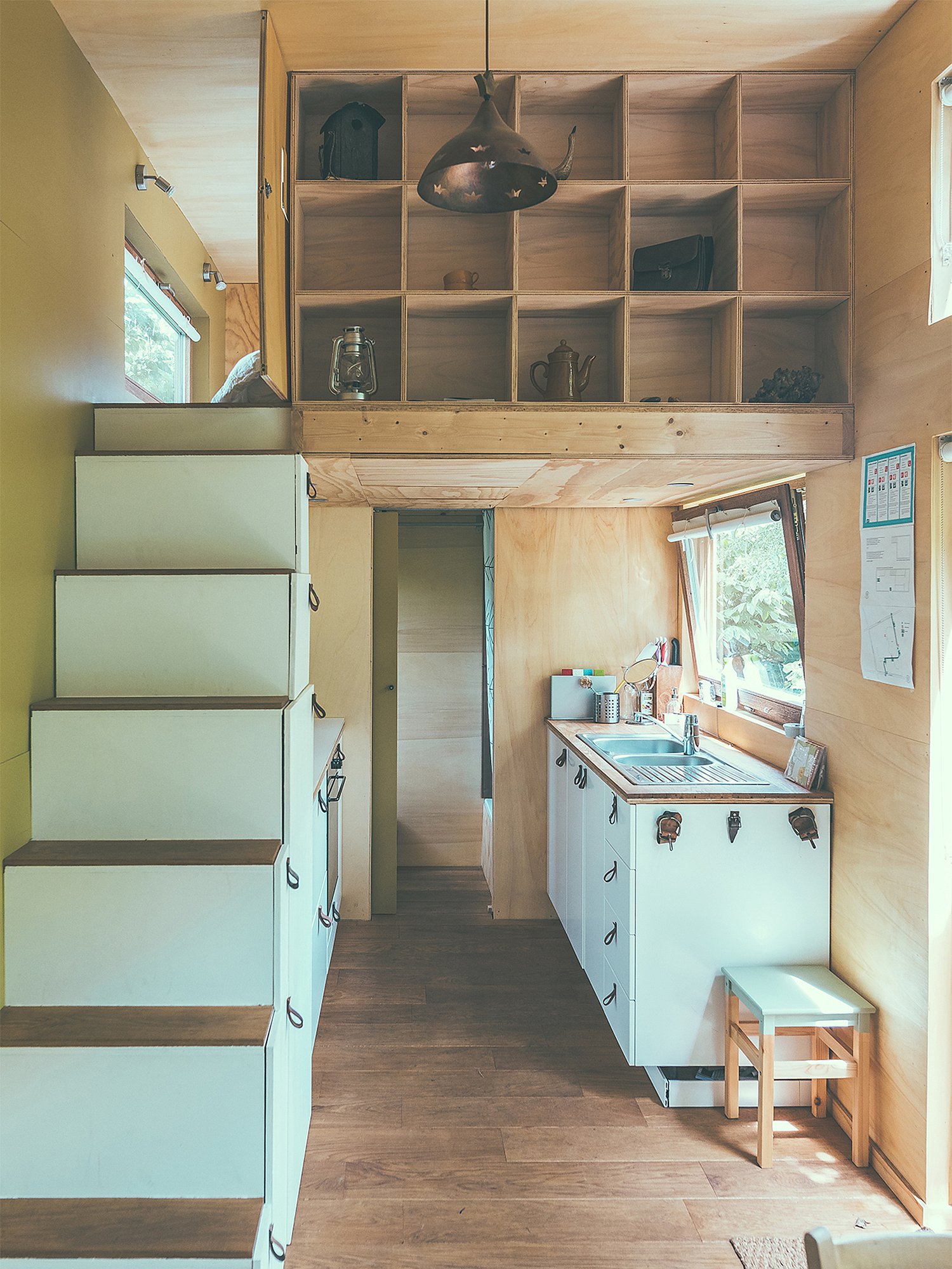 “Interior of a compact wooden pop-up hotel cabin with storage stairs, a small kitchenette, open shelving and a loft bed.
