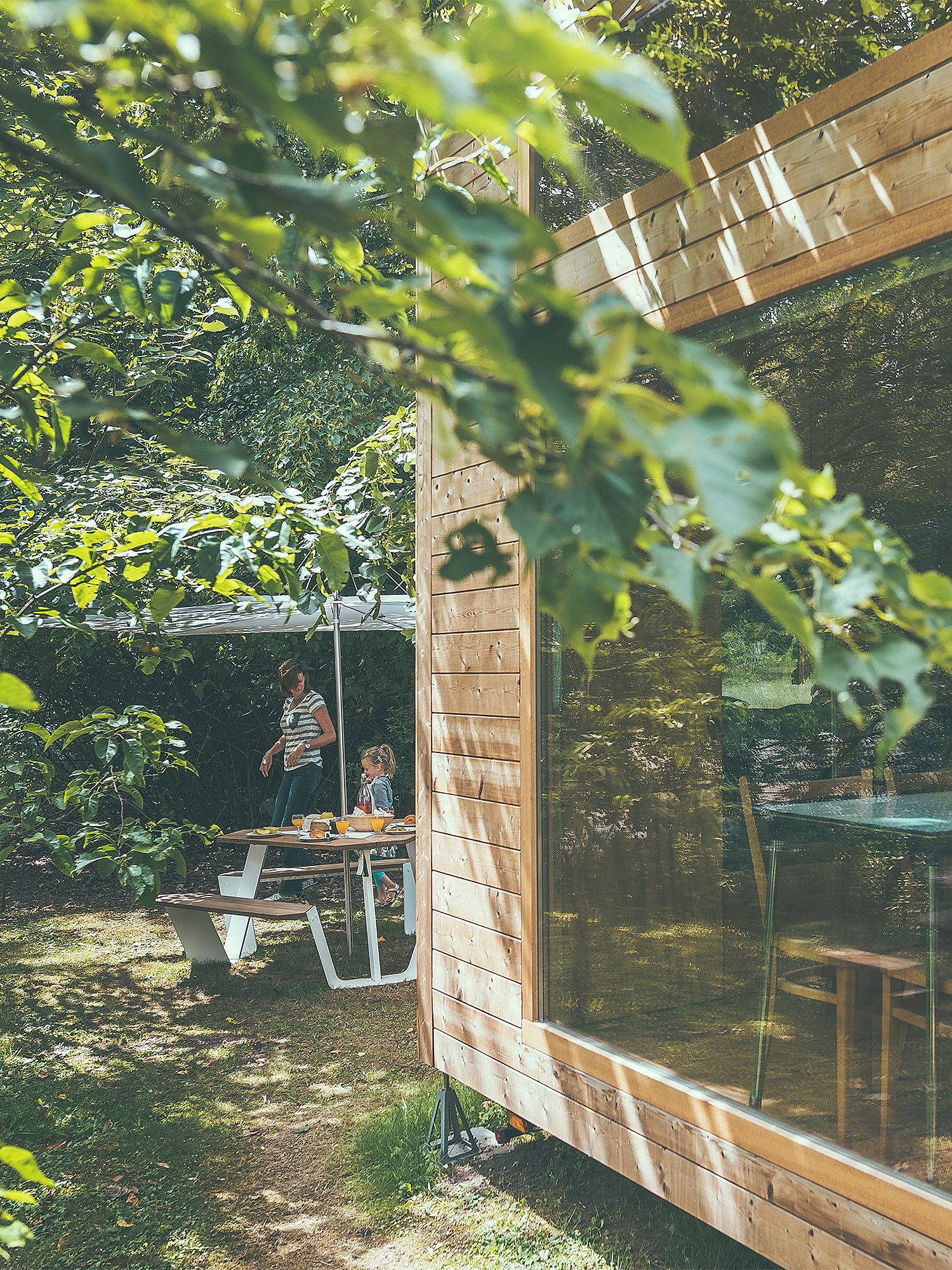 A timber pop-up hotel cabin sits among trees, with an Anker picnic table and white Inumbrina parasol set up outside in dappled sunlight.