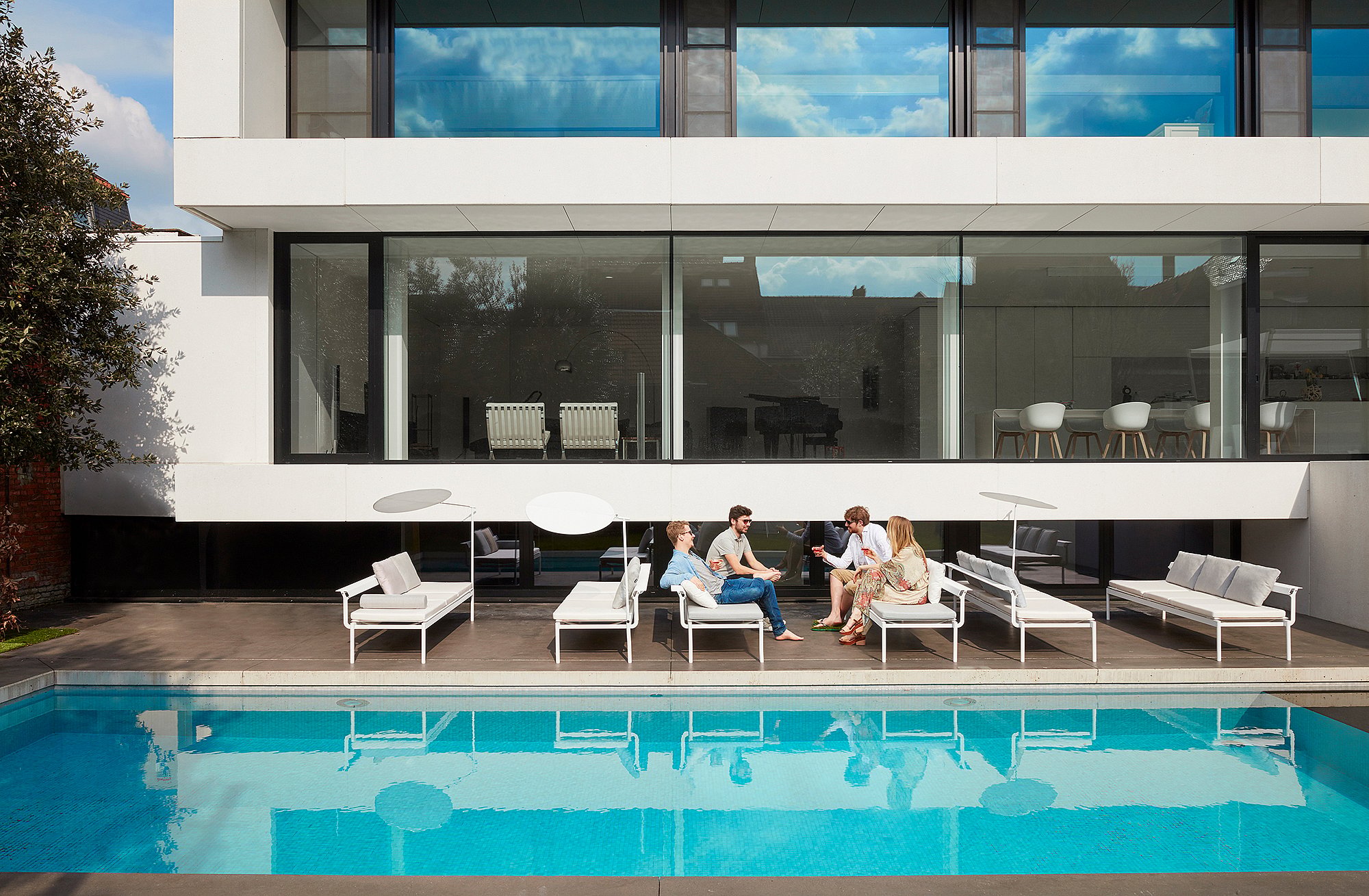   Vier Personen entspannen und unterhalten sich auf weißen Sol+Luna-Sofa-Sonnenliegen am Pool einer modernen Terrasse im Stadtzentrum. Das Haus spiegelt sich im Wasser.