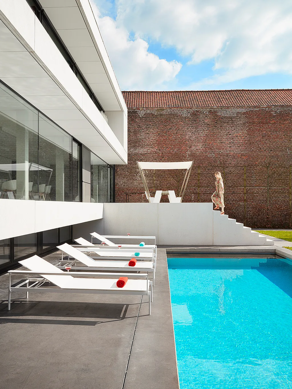White Sol+Luna sofa sunbeds line the edge of a turquoise pool beside a modern white-and-glass house, with a woman walking down the steps in the background.