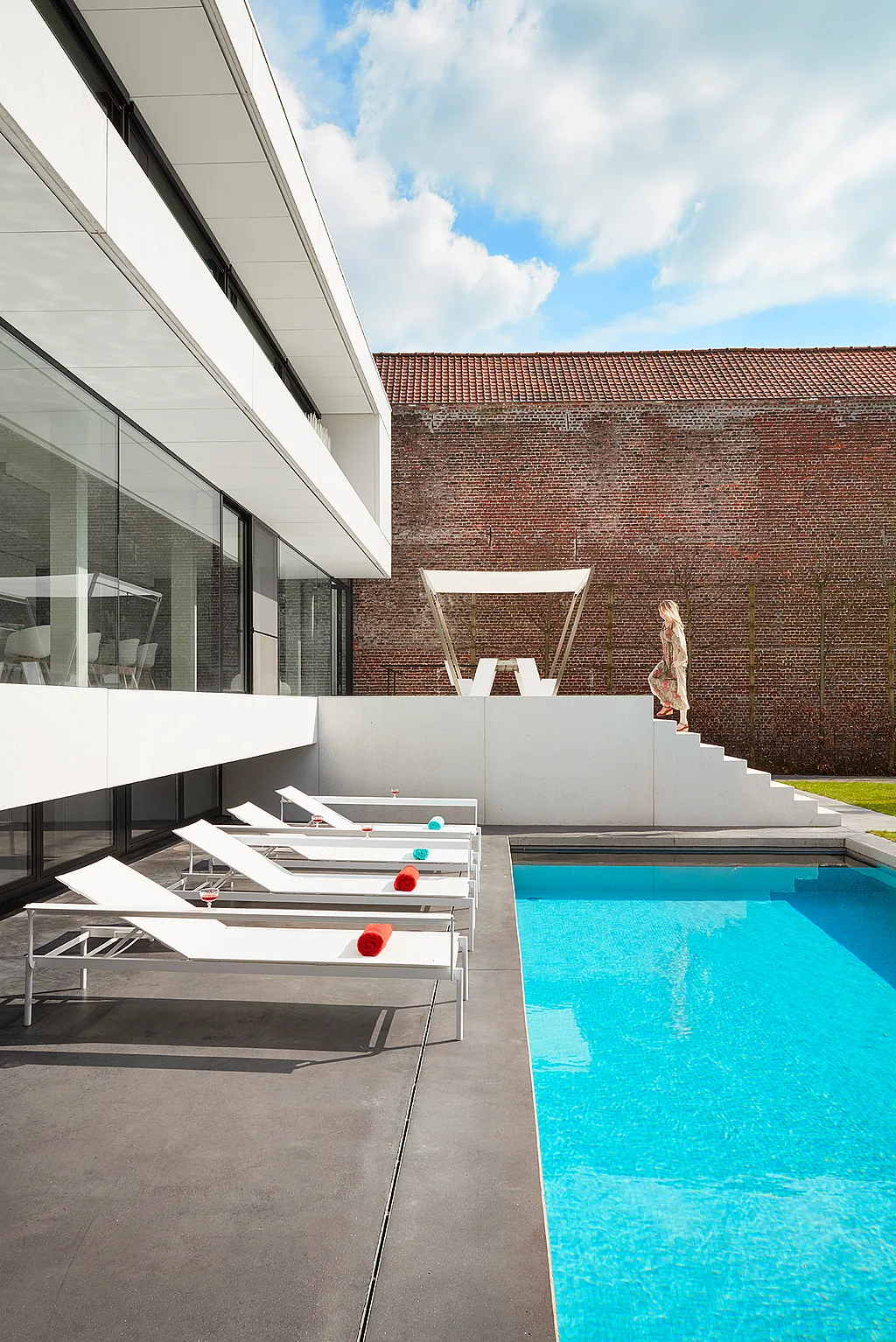 Des transats blancs Sol+Luna bordent une piscine turquoise, à côté d'une maison moderne blanche et vitrée. Une femme descend les marches en arrière-plan.