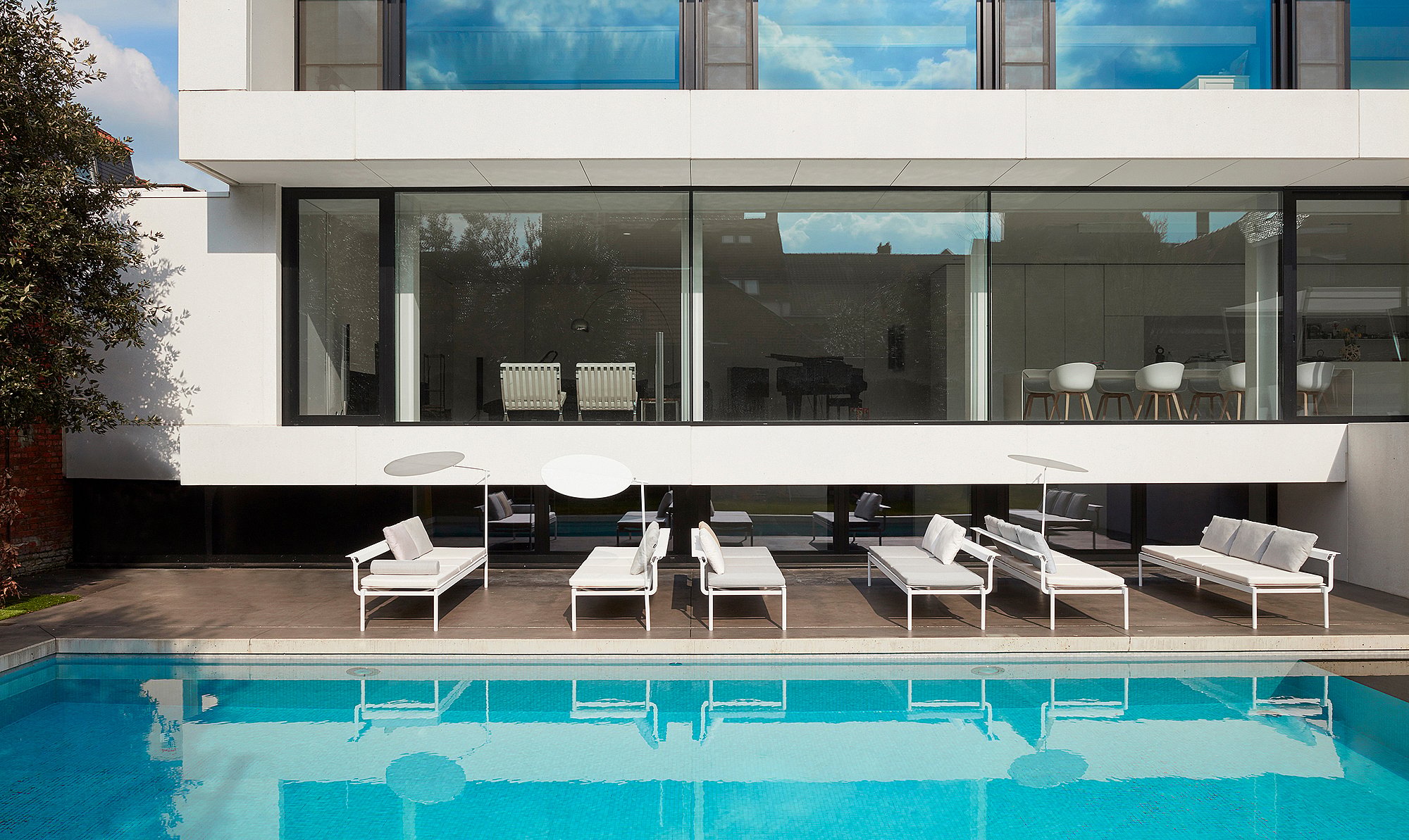   Eine Reihe weißer Sol+Luna-Sofa-Sonnenliegen steht neben einem langen Pool vor einer eleganten Glasfassade. Schmale, runde Sonnenschirme werfen sanfte Schatten.