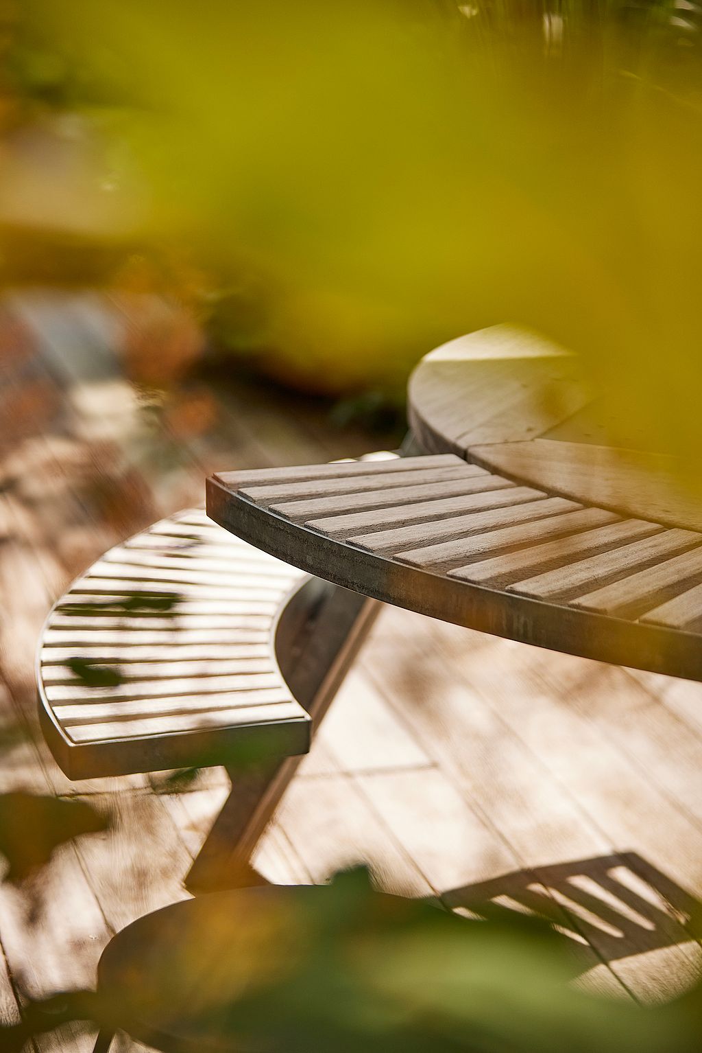 Soft-focus close-up of the curved slatted bench seats and round tabletop, with sunlight and foliage filtering across the wooden deck.