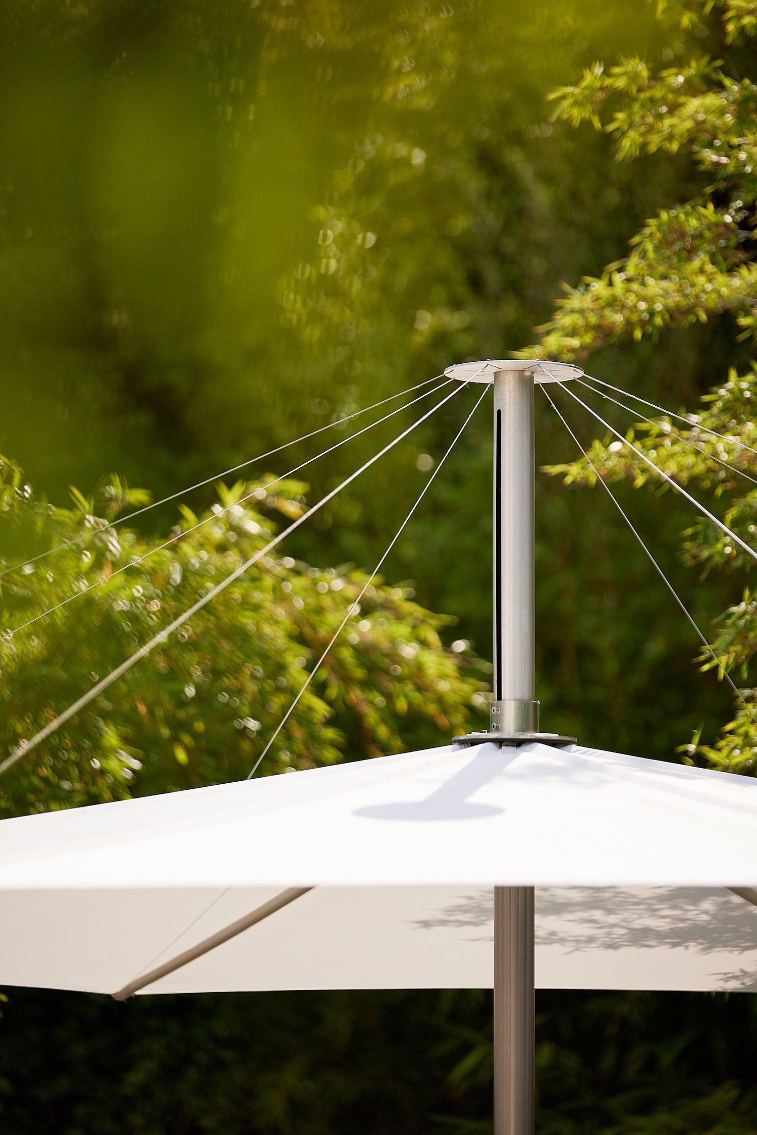 Close-up of the white inumbra parasol canopy and central pole, with tension cables extending outward against a backdrop of greenery.
