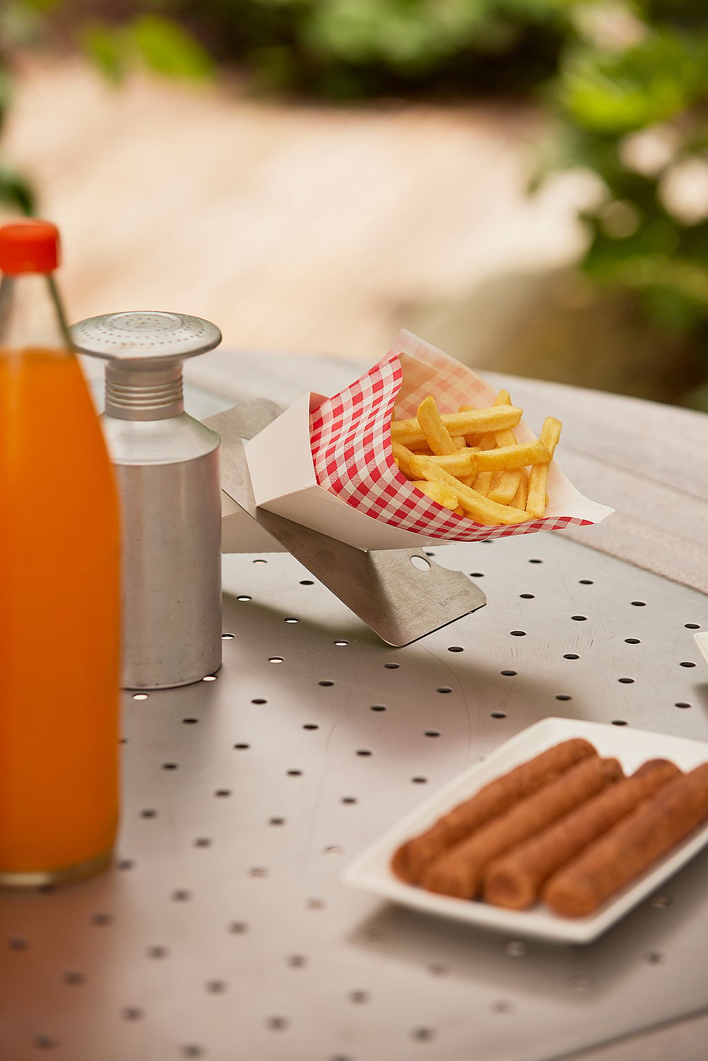 Close-up of a perforated tabletop with a paper tray of fries, a metal shaker, an orange drink bottle, and a plate of sausages.