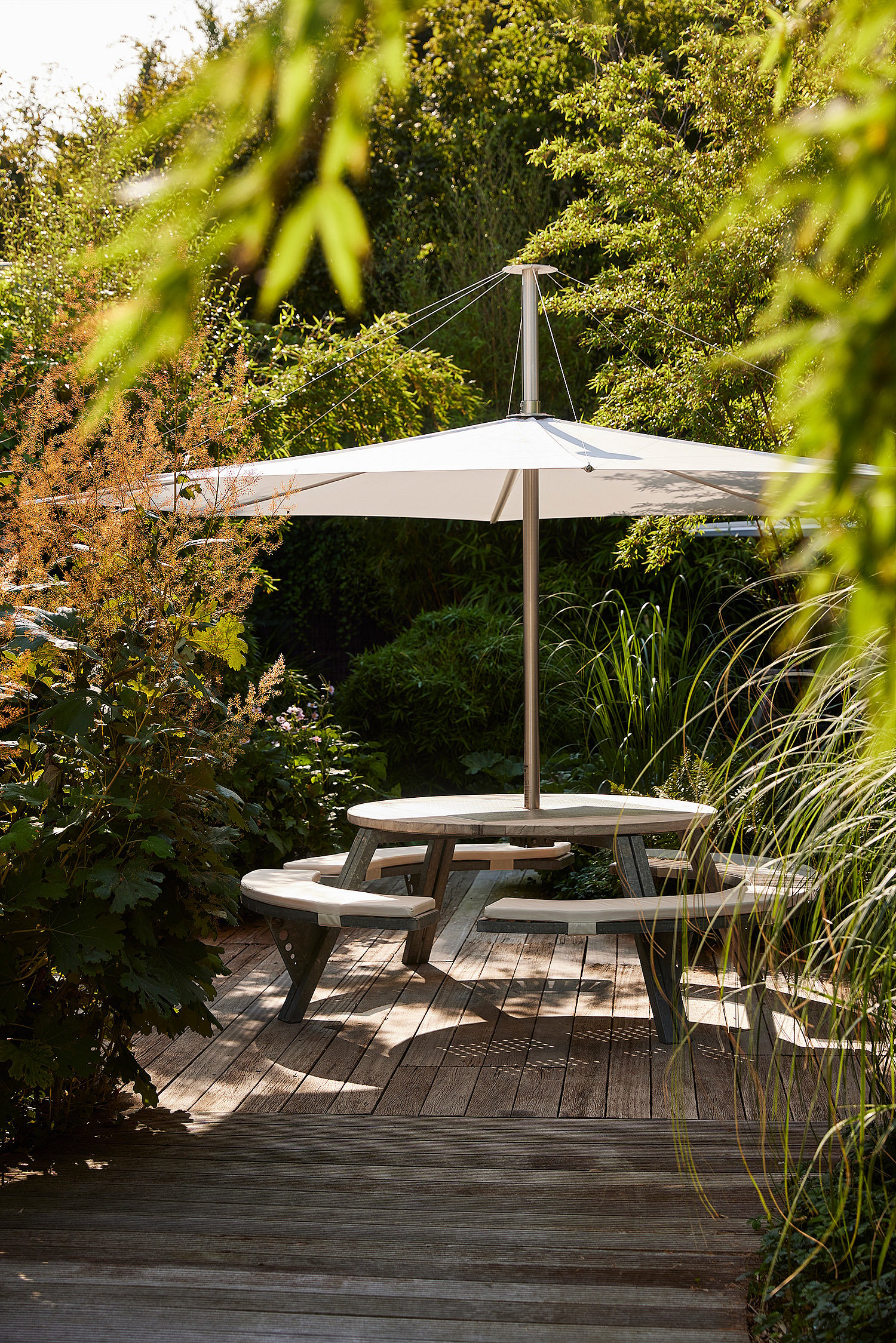 Een ronde grijze Gargantua-tafel met een witte Inumbra parasol op een houten terras, omringd door tuingroen.