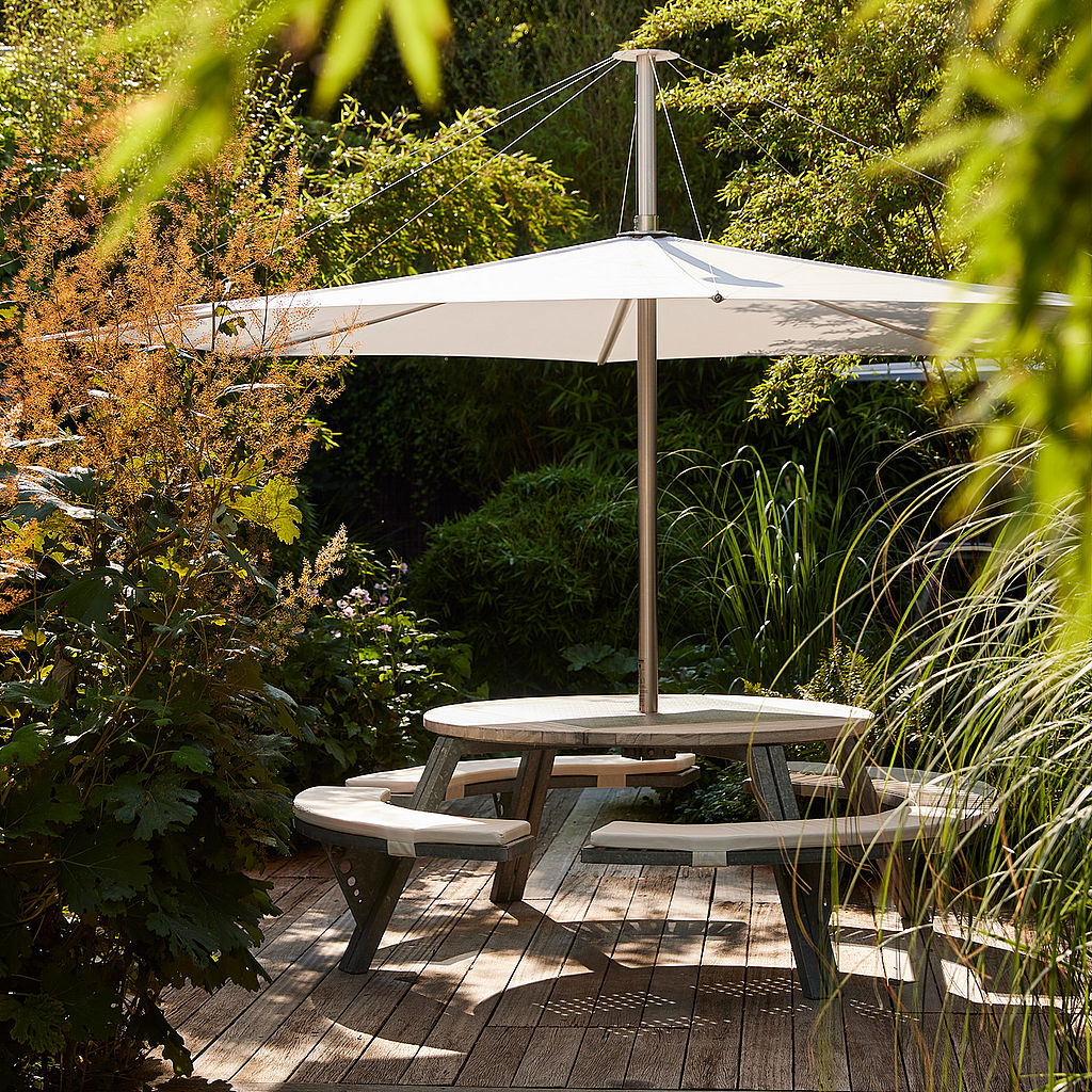 Een ronde grijze Gargantua-tafel met een witte Inumbra parasol op een houten terras, omringd door tuingroen.