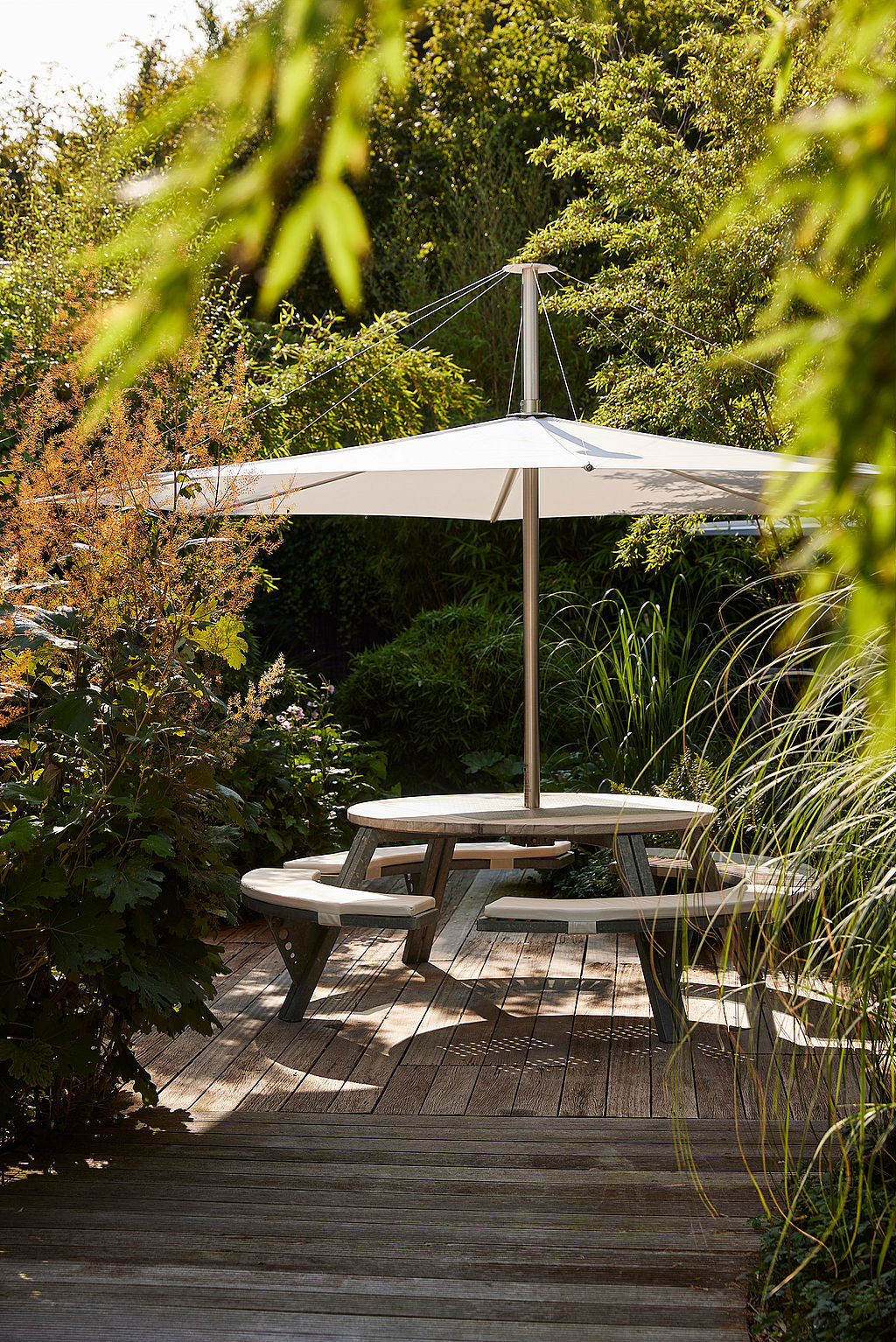 Round gargantua picnic table under a white inumbra parasol on a wooden terrace, framed by leafy branches and surrounded by lush green planting.”