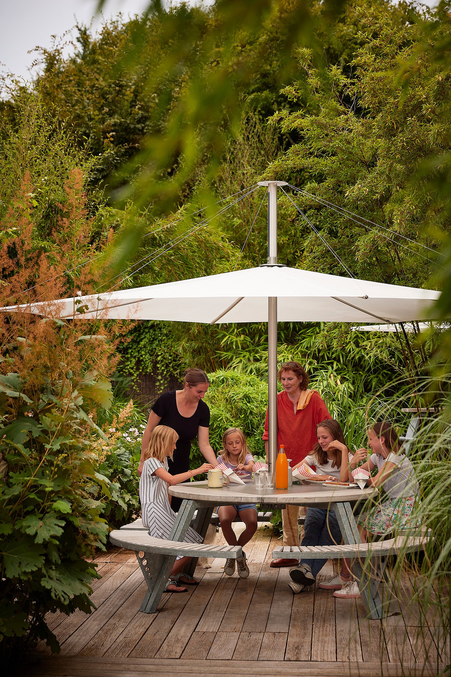 Group of adults and children gathered around the round gargantua picnic table under a white parasol, eating and talking in a lush garden setting.