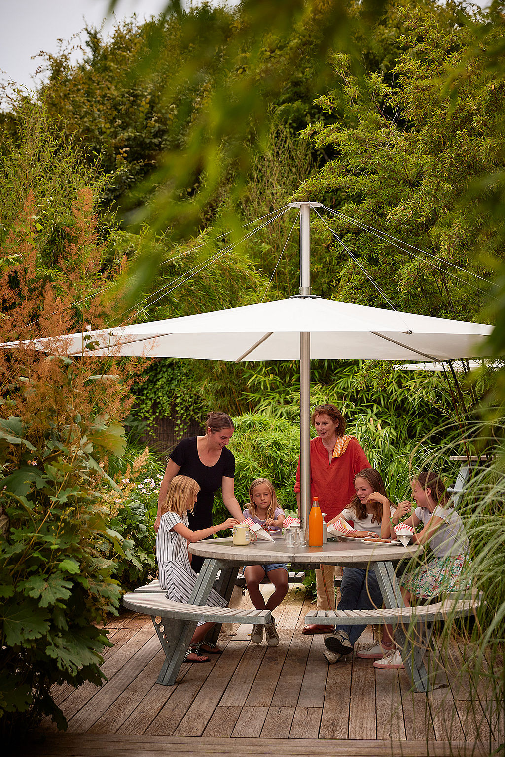 Group of adults and children gathered around the round gargantua picnic table under a white parasol, eating and talking in a lush garden setting.