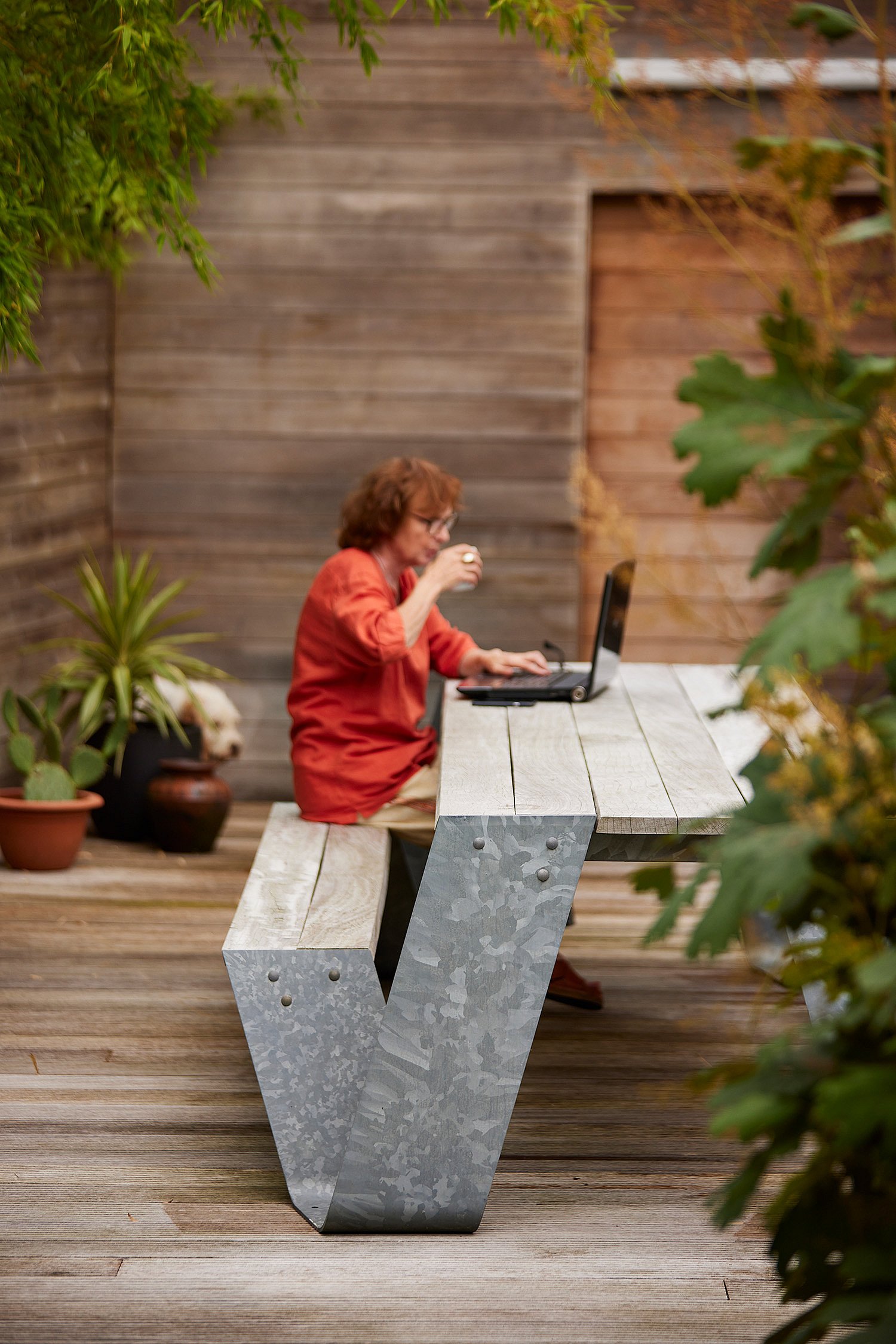 Side view of a woman drinking from a cup while working on a laptop at the picnic table on a wooden deck, framed by potted plants and a wood-clad wall.