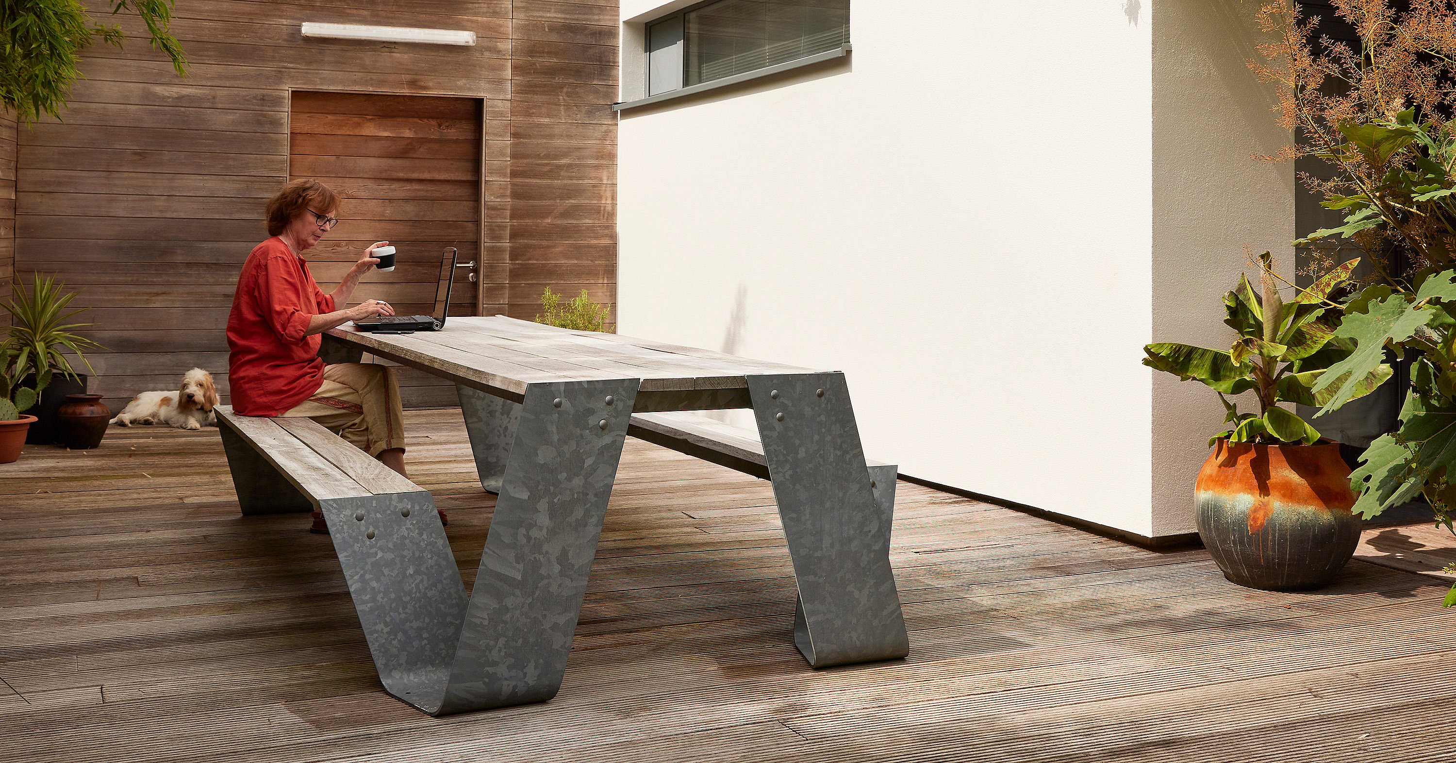 Woman working on a laptop at a weathered wooden hopper picnic table with galvanized legs on a sunlit deck, with a dog resting near the wall.