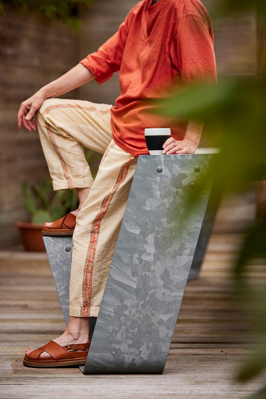 Close-up of a seated person in an orange top leaning against the galvanized table leg while holding a reusable cup.