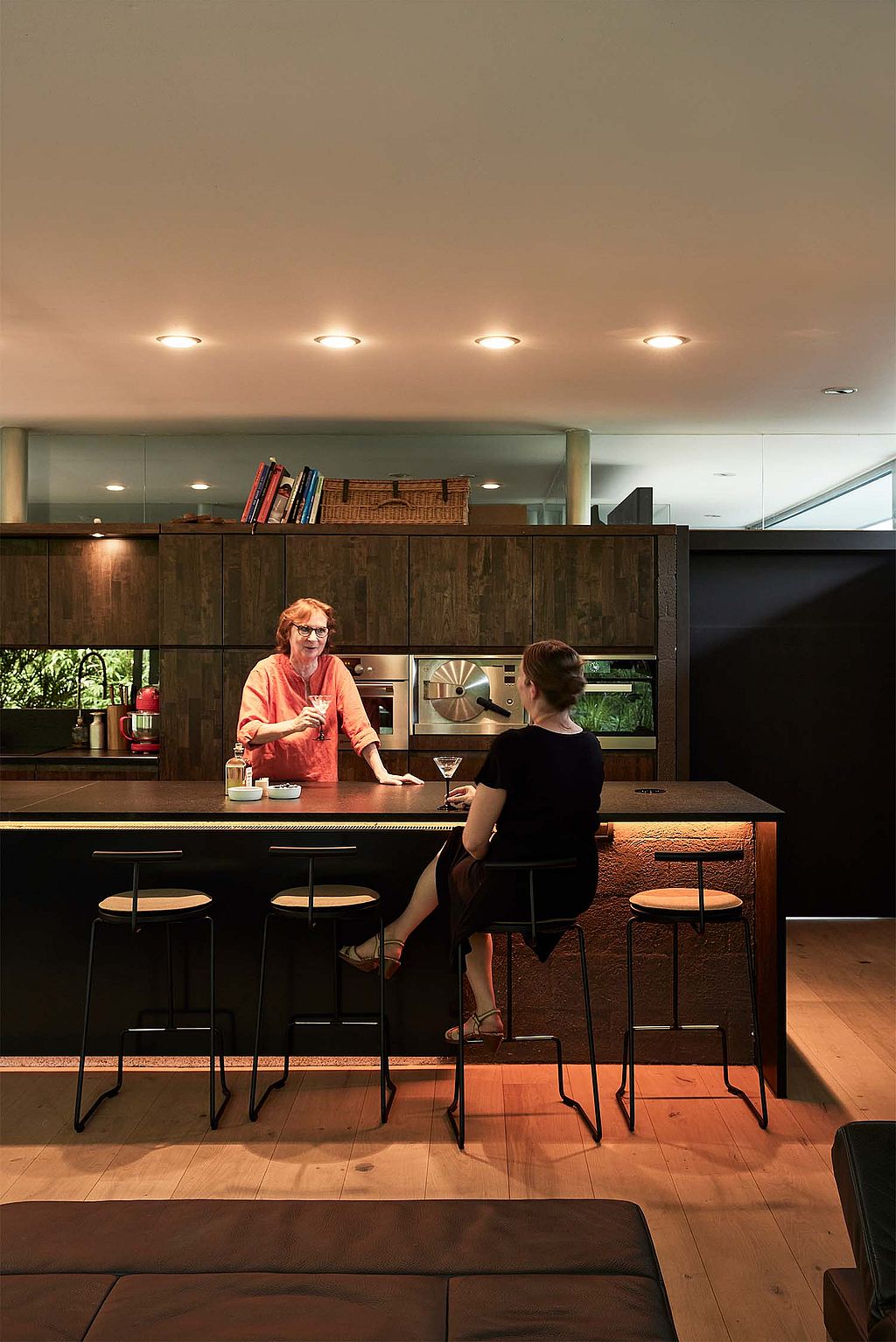 Two women share drinks at a dark kitchen island with tall black tiki stools in a warmly lit modern interior.