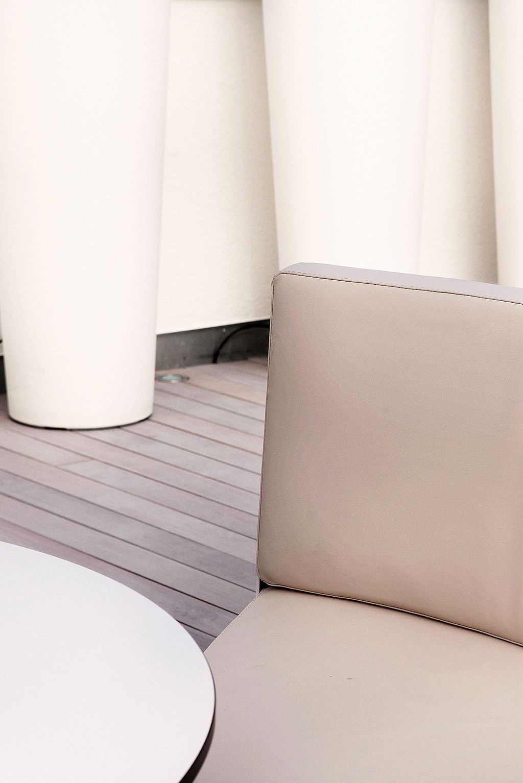 Close-up of a beige seat and backrest beside a round white tabletop, with tall white cylindrical planters in the background.