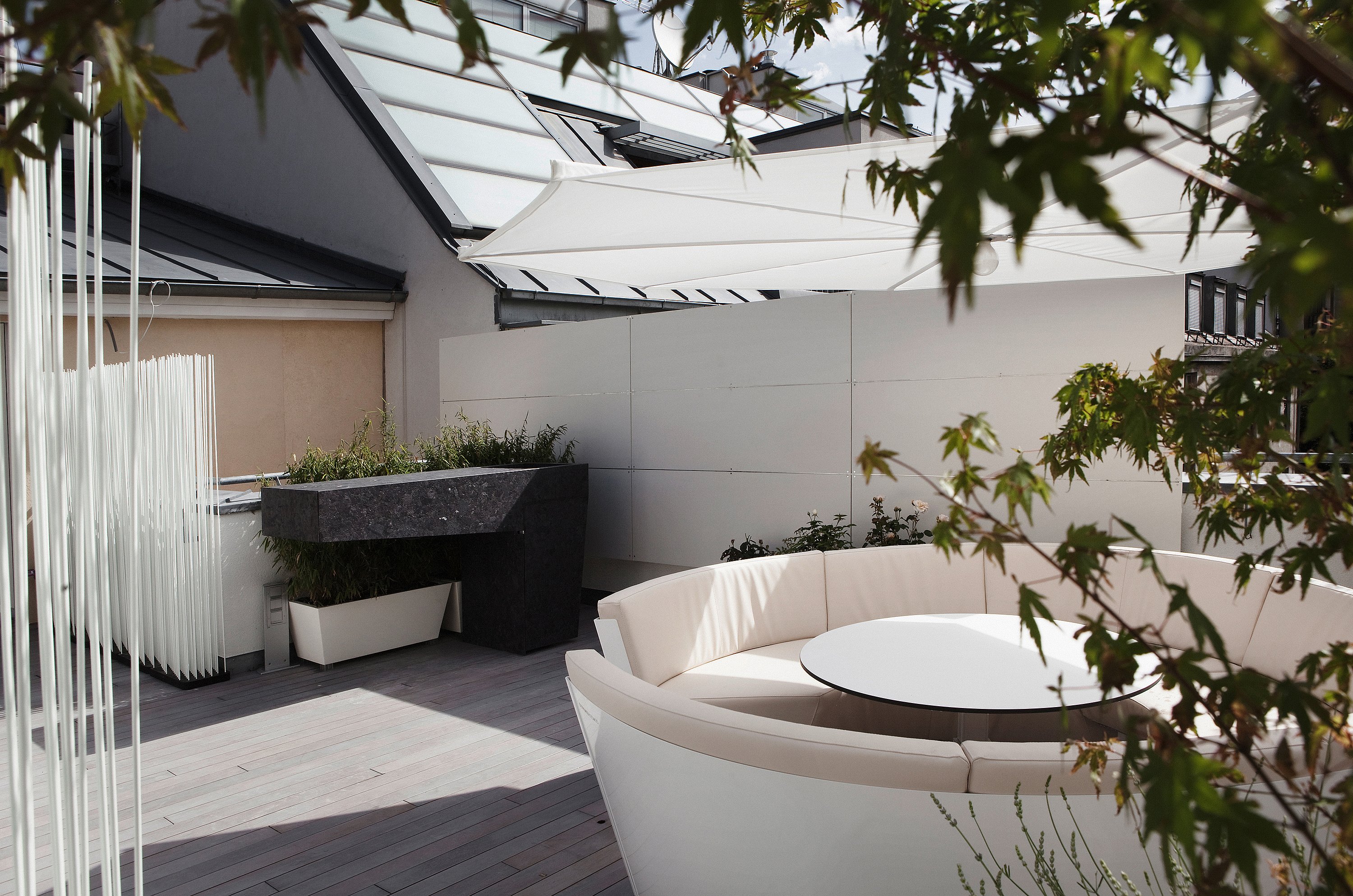 Curved white outdoor lounge with a round white table under a large white parasol on a timber deck, partly framed by leafy branches and rooftop walls.
