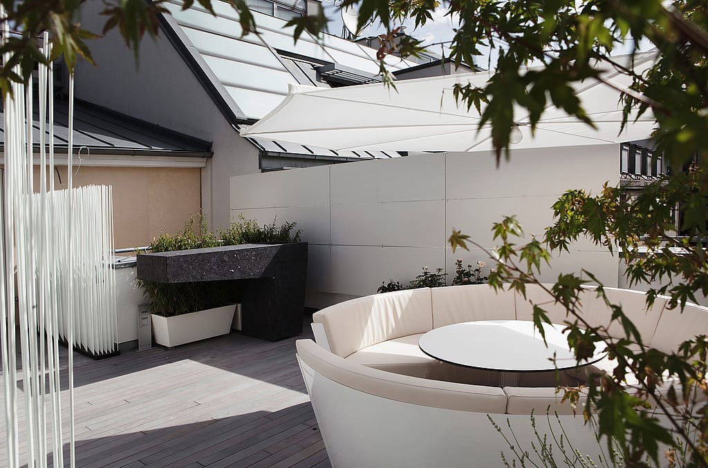 Curved white outdoor lounge with a round white table under a large white parasol on a timber deck, partly framed by leafy branches and rooftop walls.