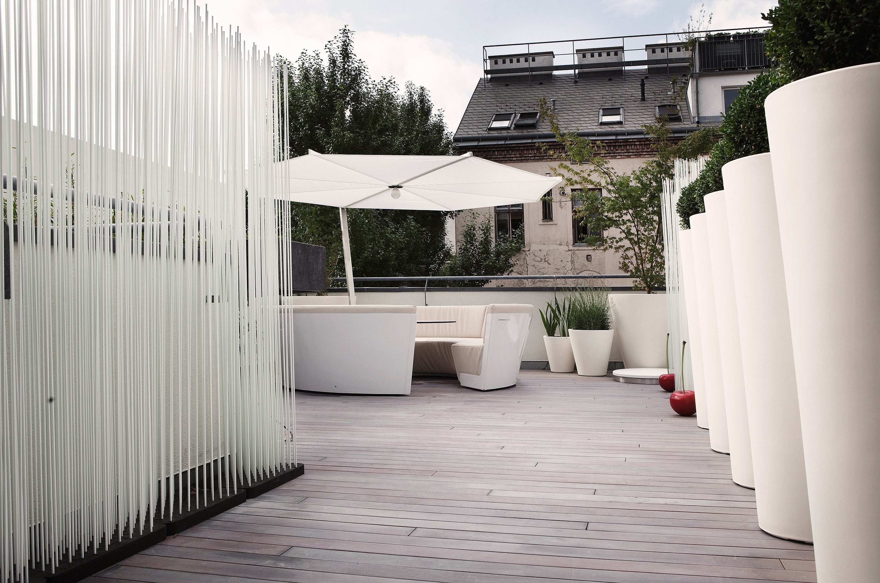 Weiße Dachterrasse mit einer runden Kosmos-Lounge im Freien und einem runden Tisch unter einem großen weißen Sonnenschirm, eingerahmt von hohen weißen Pflanzgefäßen und einem Schirm aus vertikalen weißen Stäben.