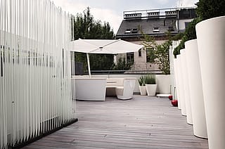 View along the timber rooftop deck toward a white circular lounge under a white parasol, with tall white planters on one side and a screen of vertical white rods on the other.