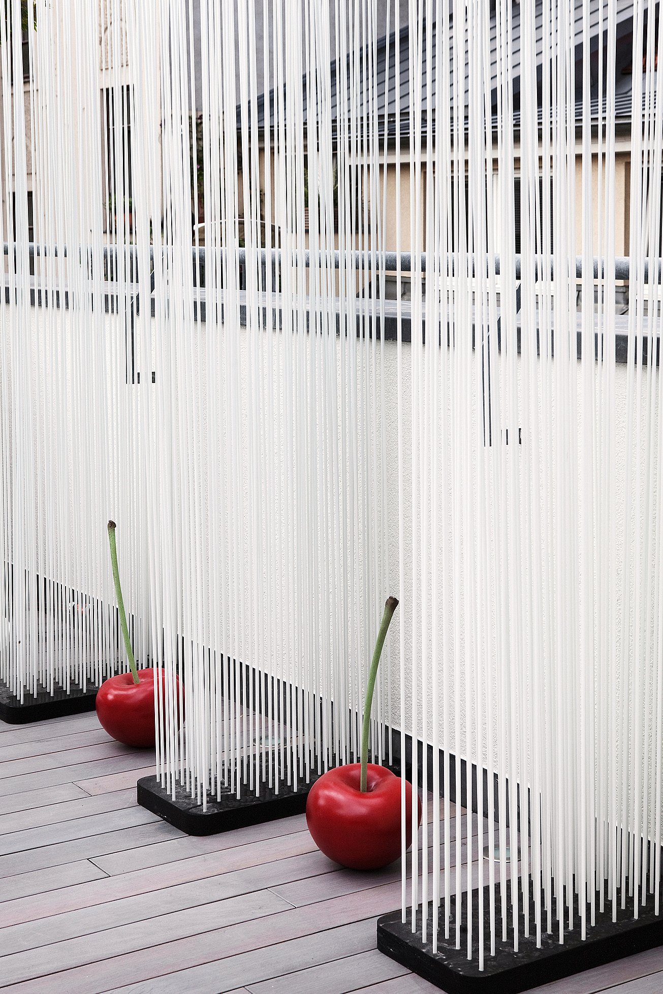 Screen made of dense vertical white rods on black bases, with two glossy red cherry ornaments placed on the timber deck in front.