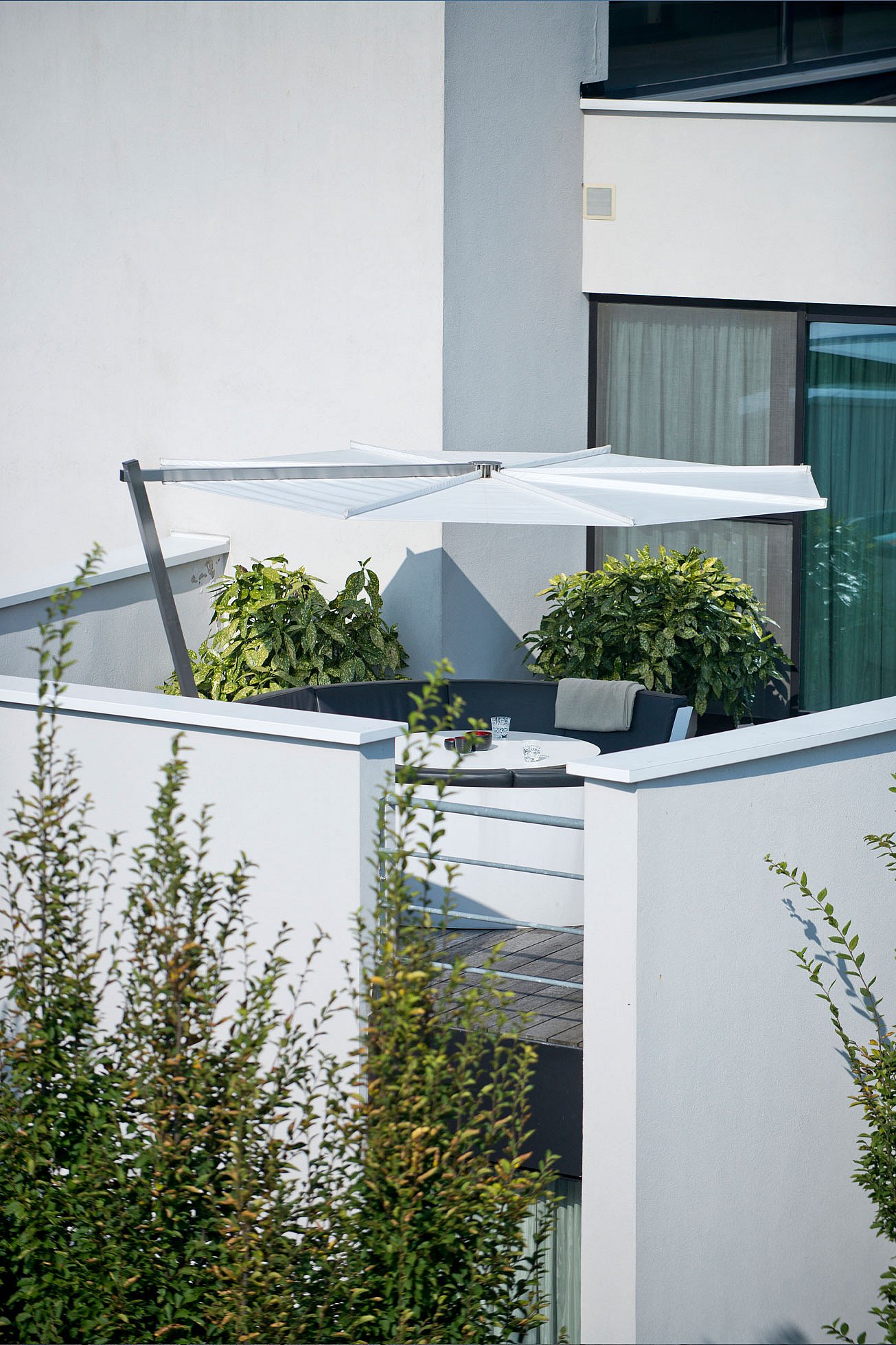 Vue lointaine d’une terrasse privée sur le toit entourée de murs blancs, montrant le salon circulaire Kosmos niché sous un parasol blanc en porte-à-faux à côté d’arbustes en pot et de grandes fenêtres.