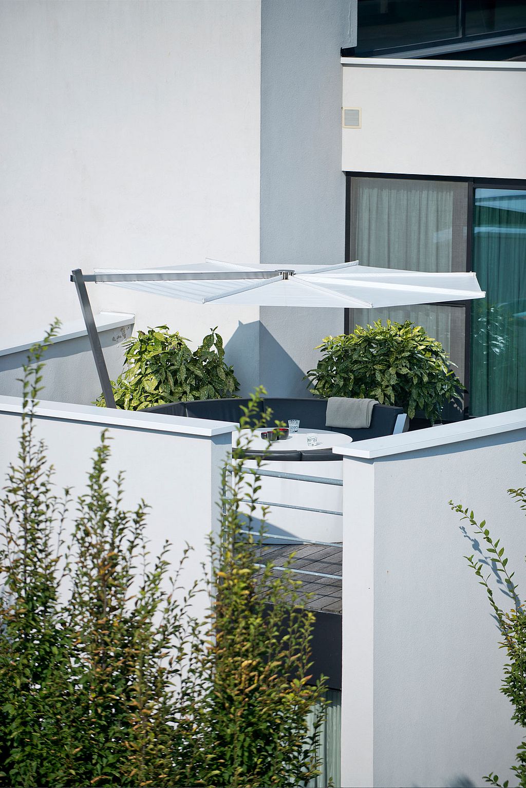 Vue lointaine d’une terrasse privée sur le toit entourée de murs blancs, montrant le salon circulaire Kosmos niché sous un parasol blanc en porte-à-faux à côté d’arbustes en pot et de grandes fenêtres.