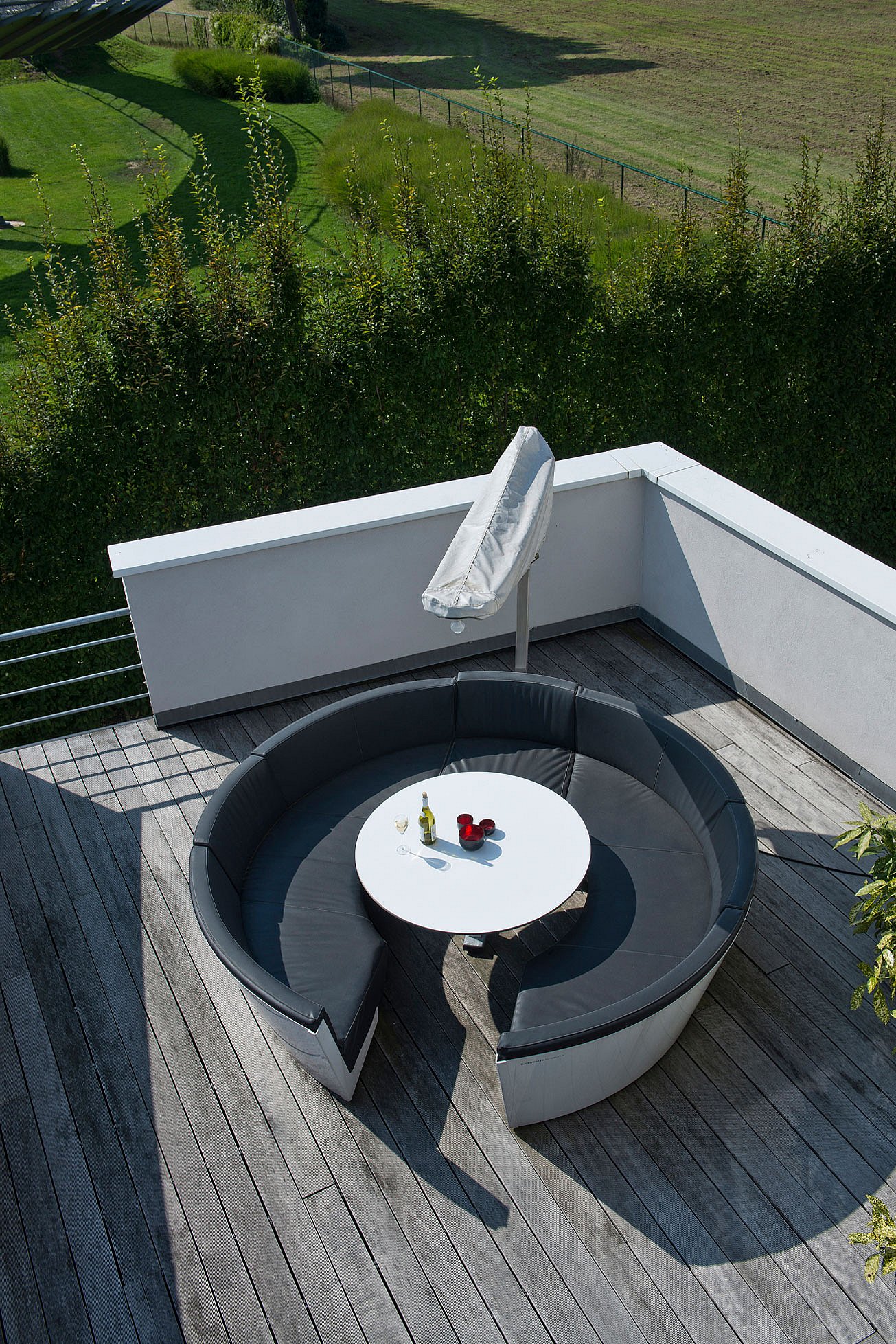 Vue de dessus d’un canapé circulaire en plein air avec des coussins sombres et une table ronde blanche sur une terrasse en bois patinée, avec le parasol plié au-dessus et des champs verts au-delà.