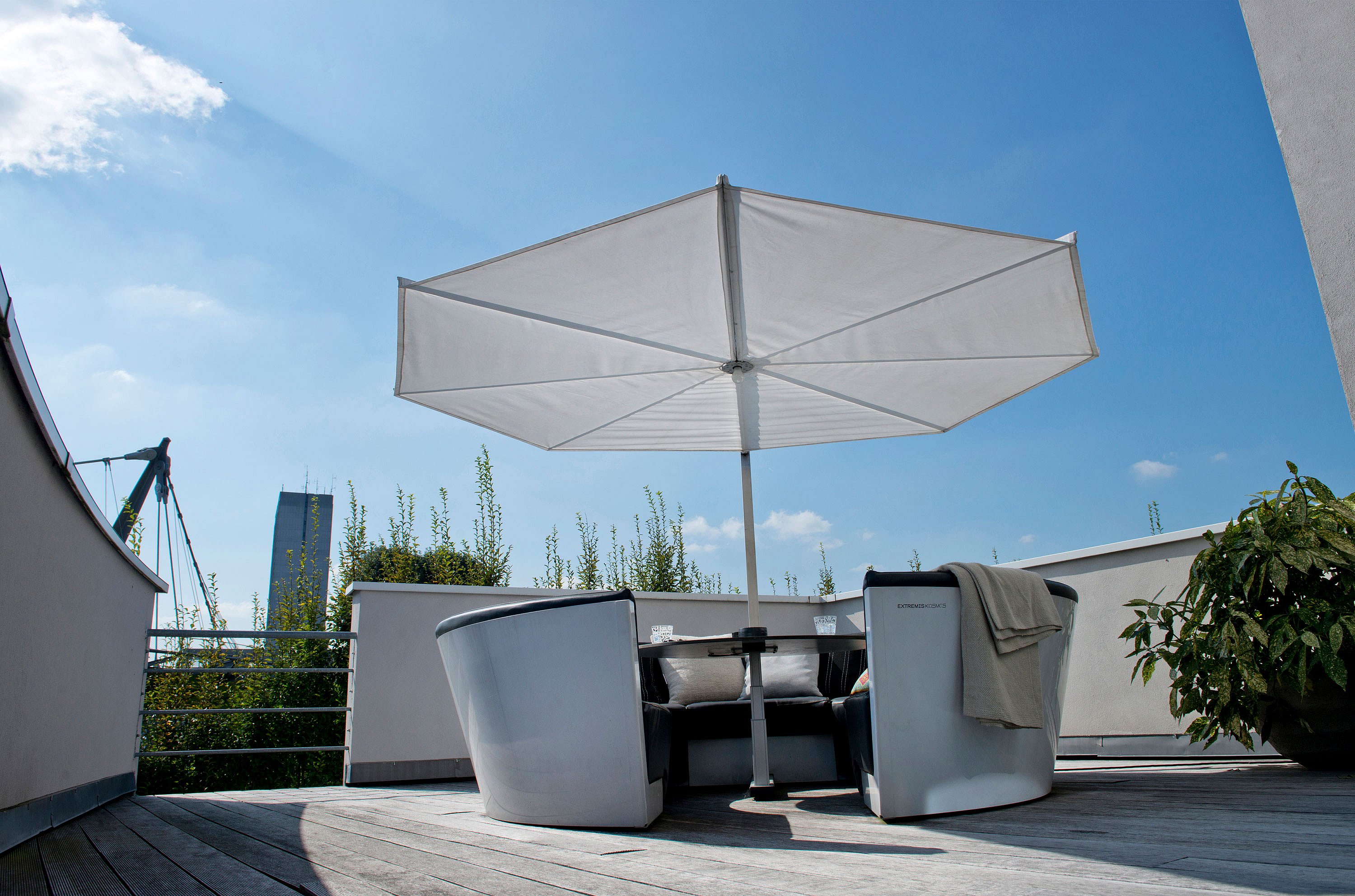 Salon circulaire en plein air avec des coussins sombres rassemblés autour d’une table ronde sous un grand parasol blanc sur une terrasse en bois sur le toit sous un ciel bleu lumineux.