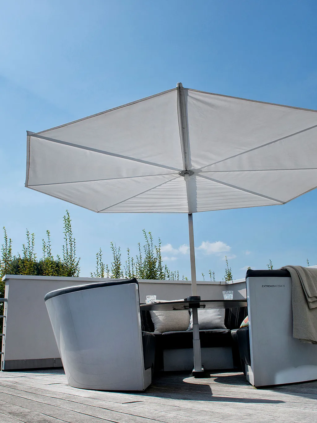 Salon circulaire en plein air avec des coussins sombres rassemblés autour d’une table ronde sous un grand parasol blanc sur une terrasse en bois sur le toit sous un ciel bleu lumineux.