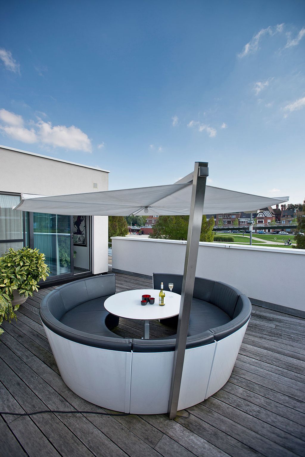 Salon circulaire en plein air avec des coussins de siège sombres et une table ronde blanche sous un parasol blanc en porte-à-faux sur une terrasse sur le toit, avec vue dégagée sur les bâtiments et la verdure à proximité.