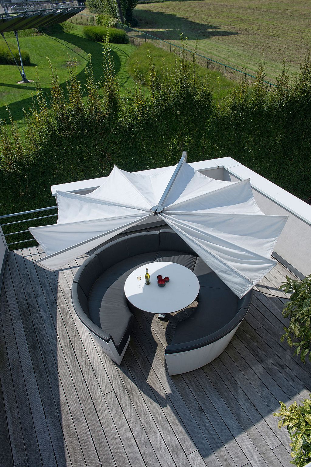 Vue aérienne d'un parasol Kosmos s'ouvrant comme des ailes au-dessus d'une assise lounge ronde grise sur une terrasse en bois.