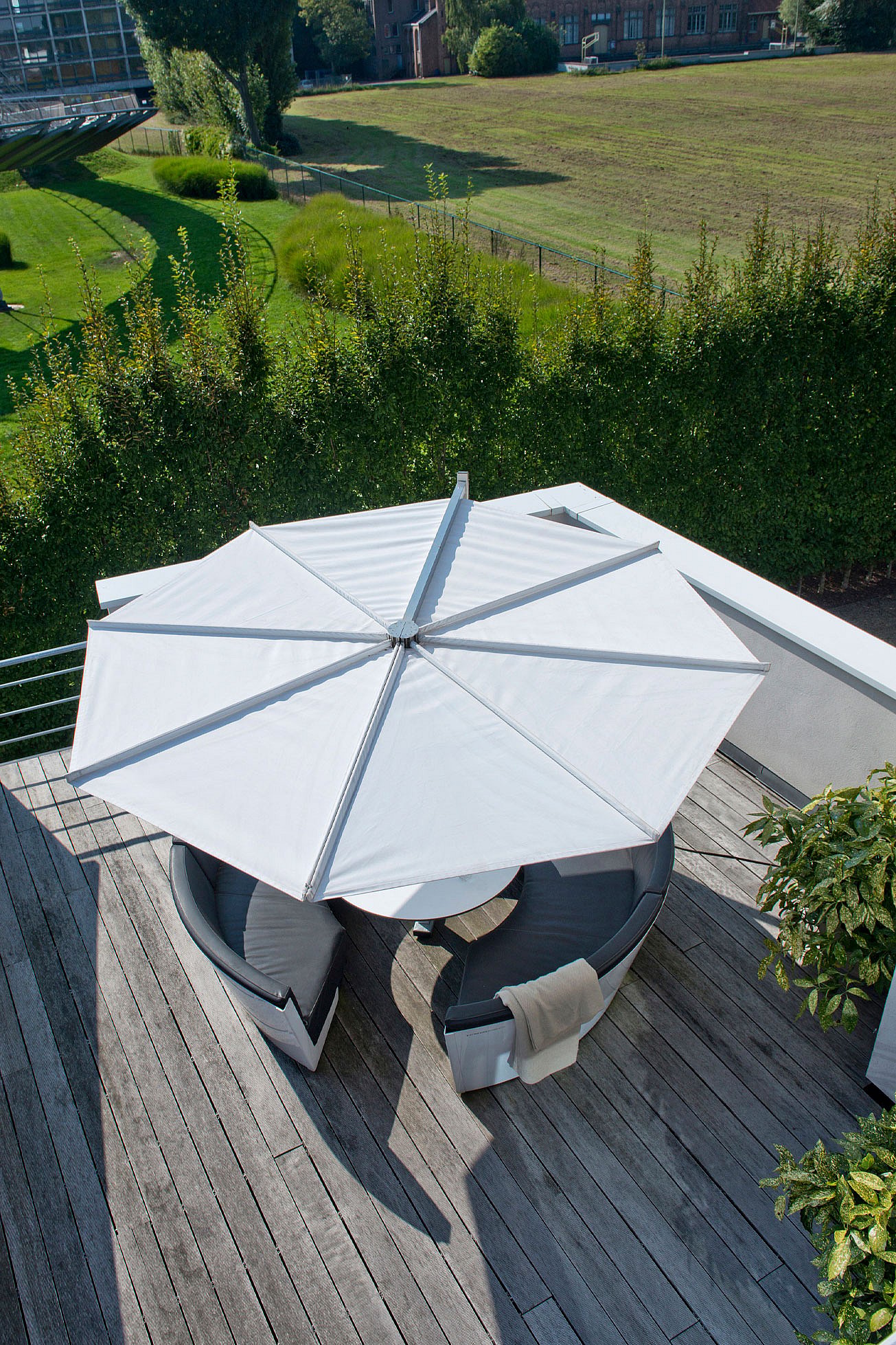 Vue aérienne du salon circulaire kosmos entièrement ombragé par un grand parasol blanc, avec un plissé drapé sur le siège et de la verdure au-delà du mur de la terrasse.