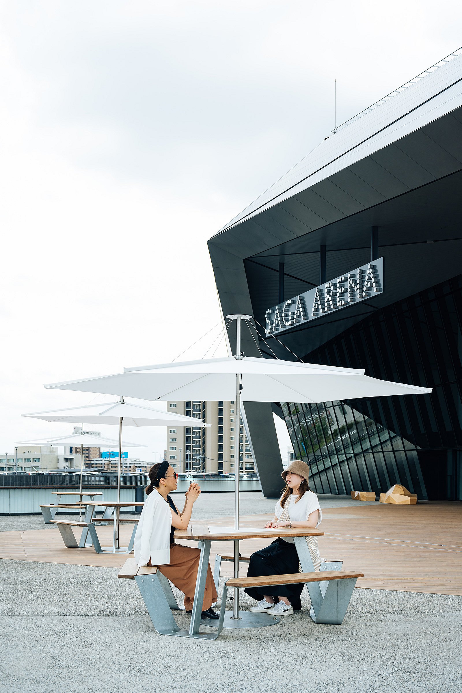 Two women seated at a anker table under a white inumbrina parasol outside the angular black façade of SAGA Arena, with more matching tables extending behind them.
