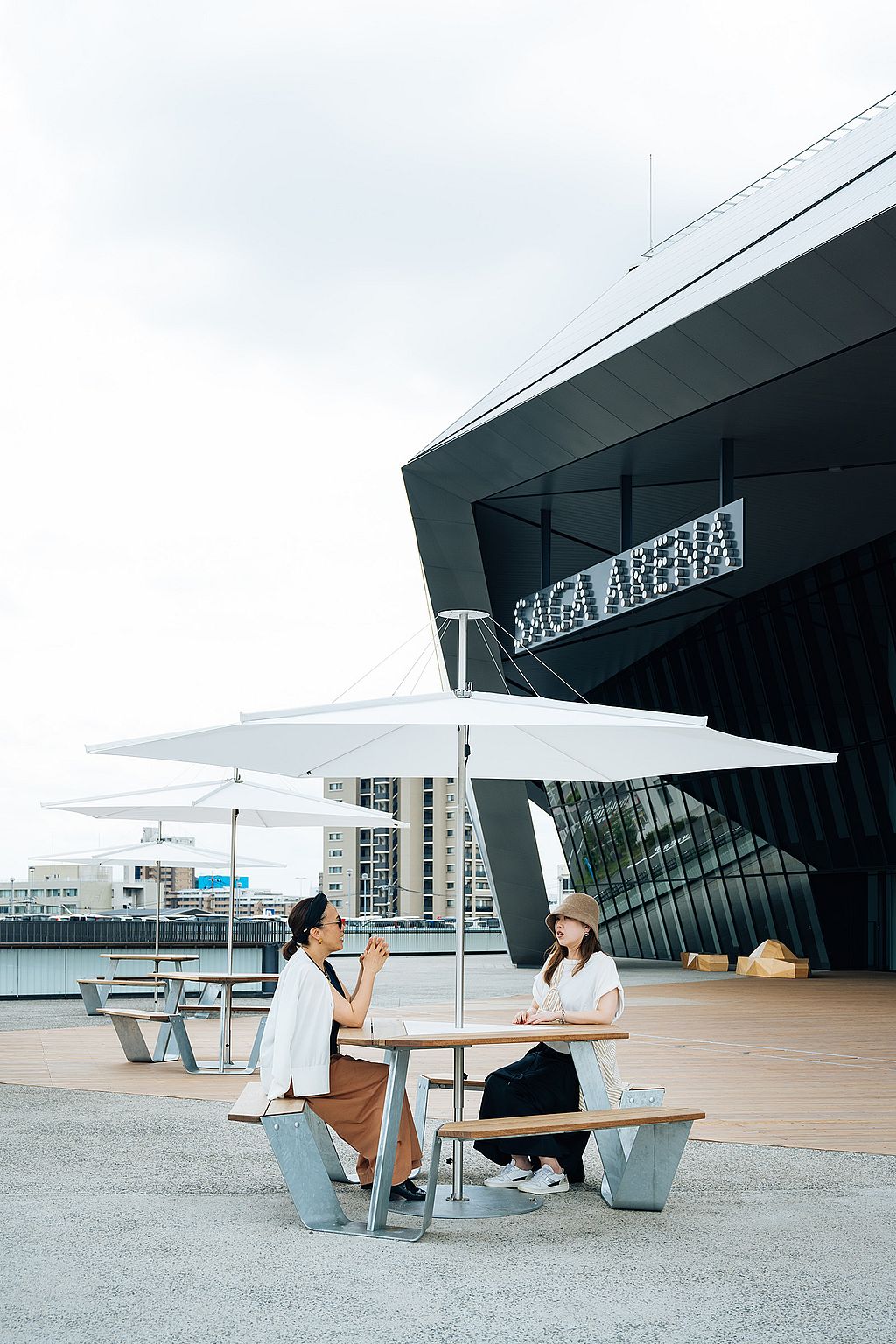 Two women seated at a anker table under a white inumbrina parasol outside the angular black façade of SAGA Arena, with more matching tables extending behind them.