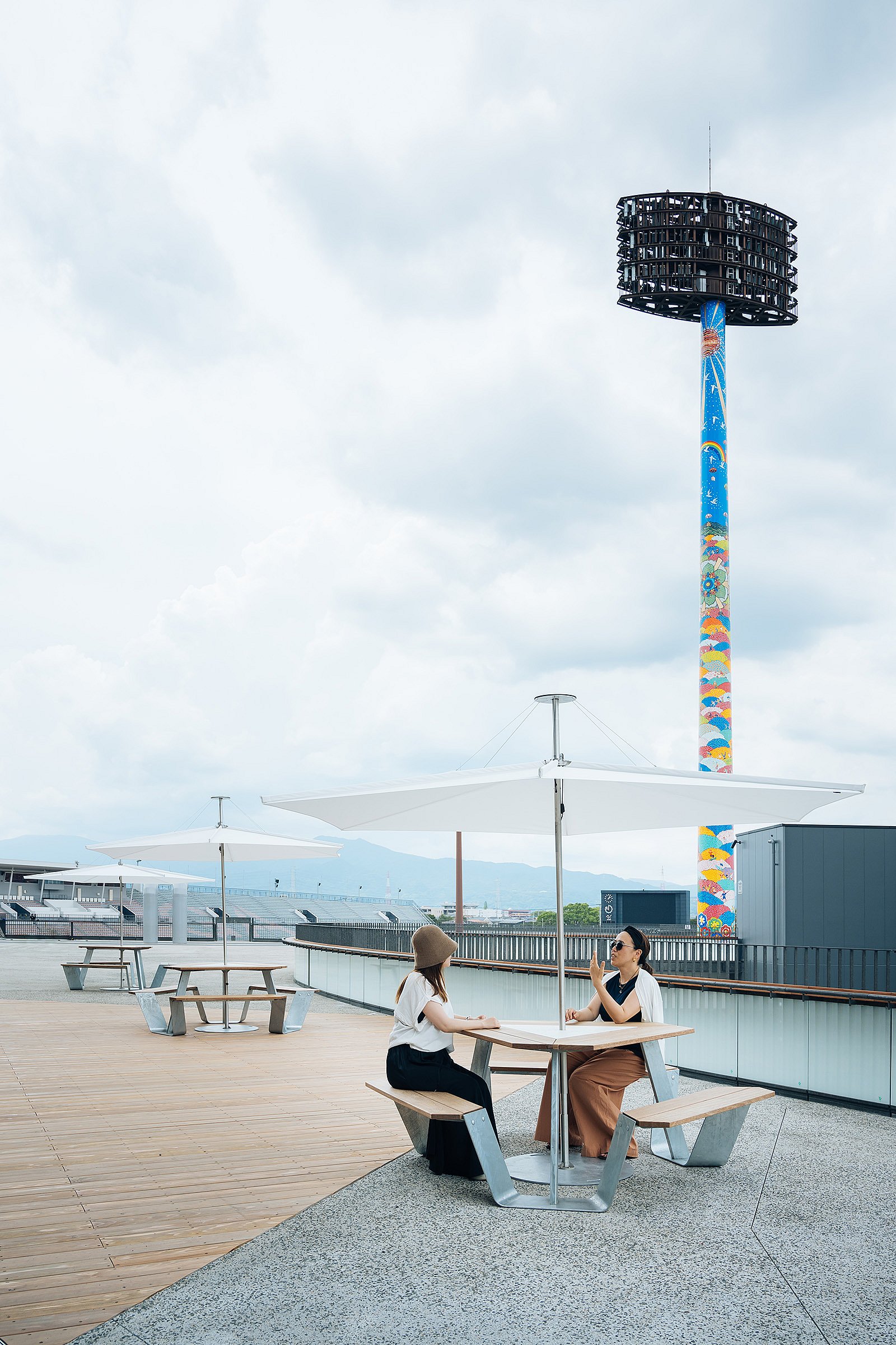 Two women sitting at a anker table under a white inumbrina parasol on an open terrace, with more inumbrina parasols, deck seating, and a tall colorful tower in the background.