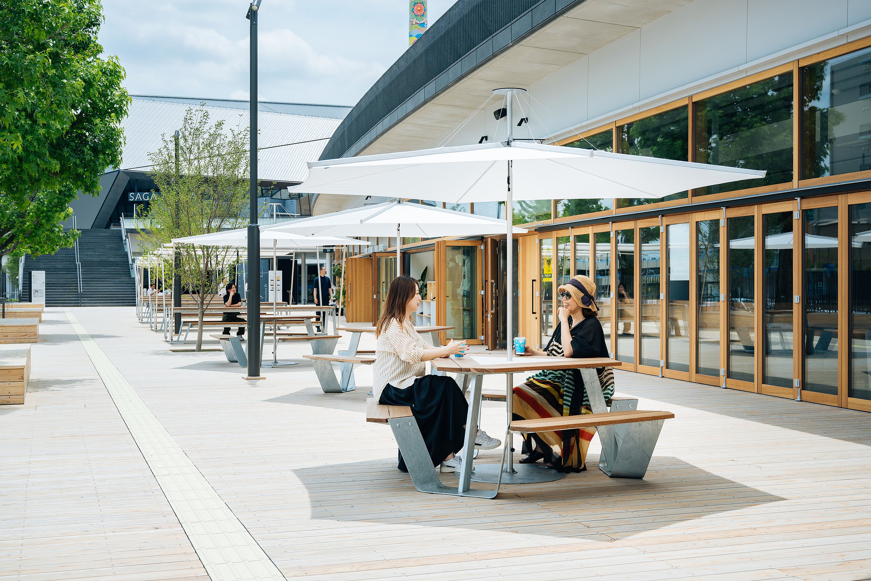Houten voetgangersdek bekleed met ankertafels en grote witte inumbrina-parasols voor een gebouw met glazen voorkant, met twee vrouwen aan de dichtstbijzijnde tafel en de arena-trappen zichtbaar op de achtergrond.