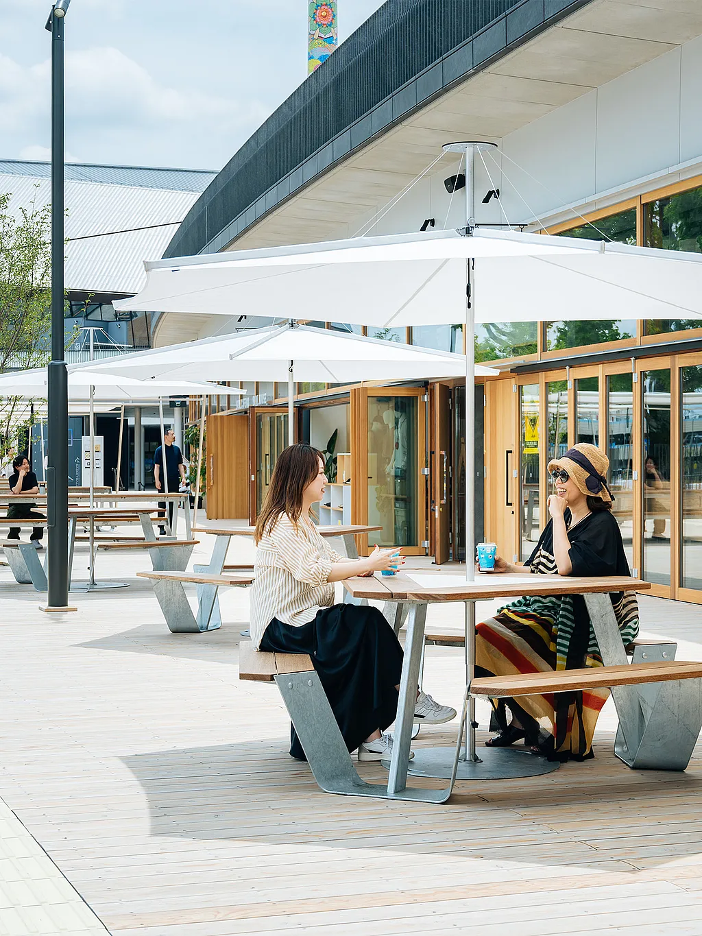 Houten voetgangersdek bekleed met ankertafels en grote witte inumbrina-parasols voor een gebouw met glazen voorkant, met twee vrouwen aan de dichtstbijzijnde tafel en de arena-trappen zichtbaar op de achtergrond.