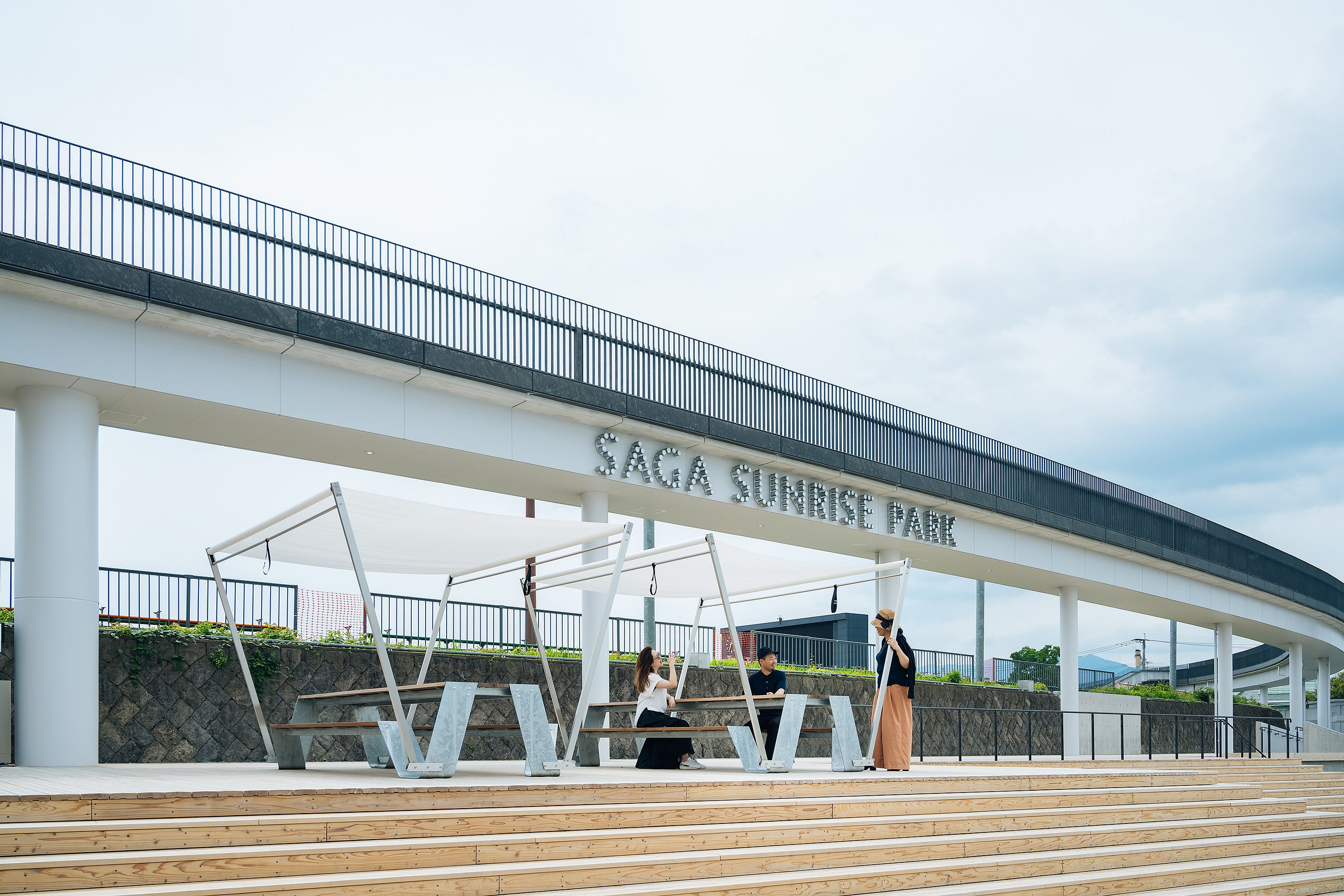 Wide stepped plaza beneath an elevated walkway signed ‘SAGA SUNRISE PARK’, with several shaded hopper picnic tables and three people gathered around one of them.