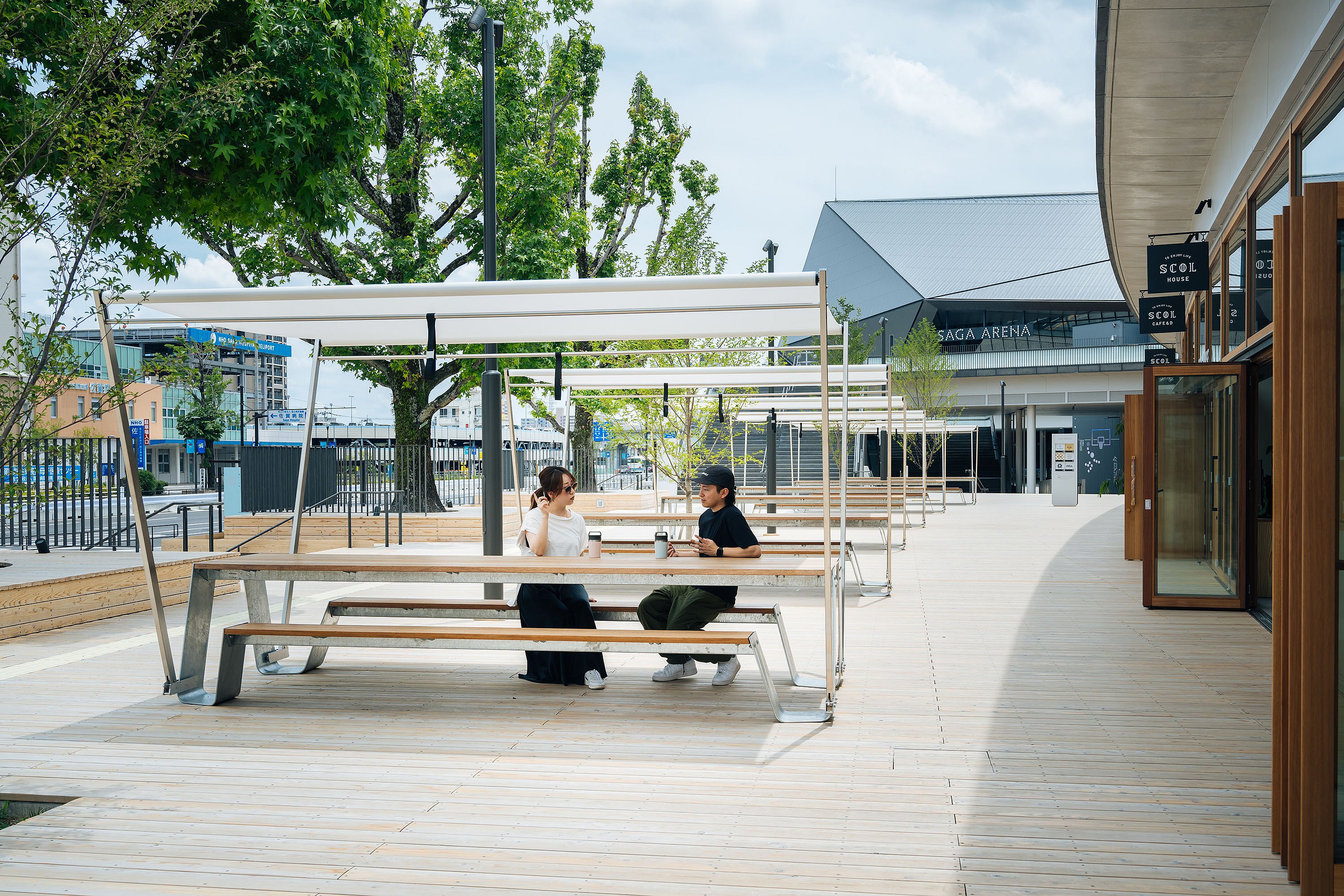 Two people seated at a long wooden hopper picnic table with integrated benches beneath a rectangular white shade canopy on a broad outdoor deck, with SAGA Arena visible behind them.