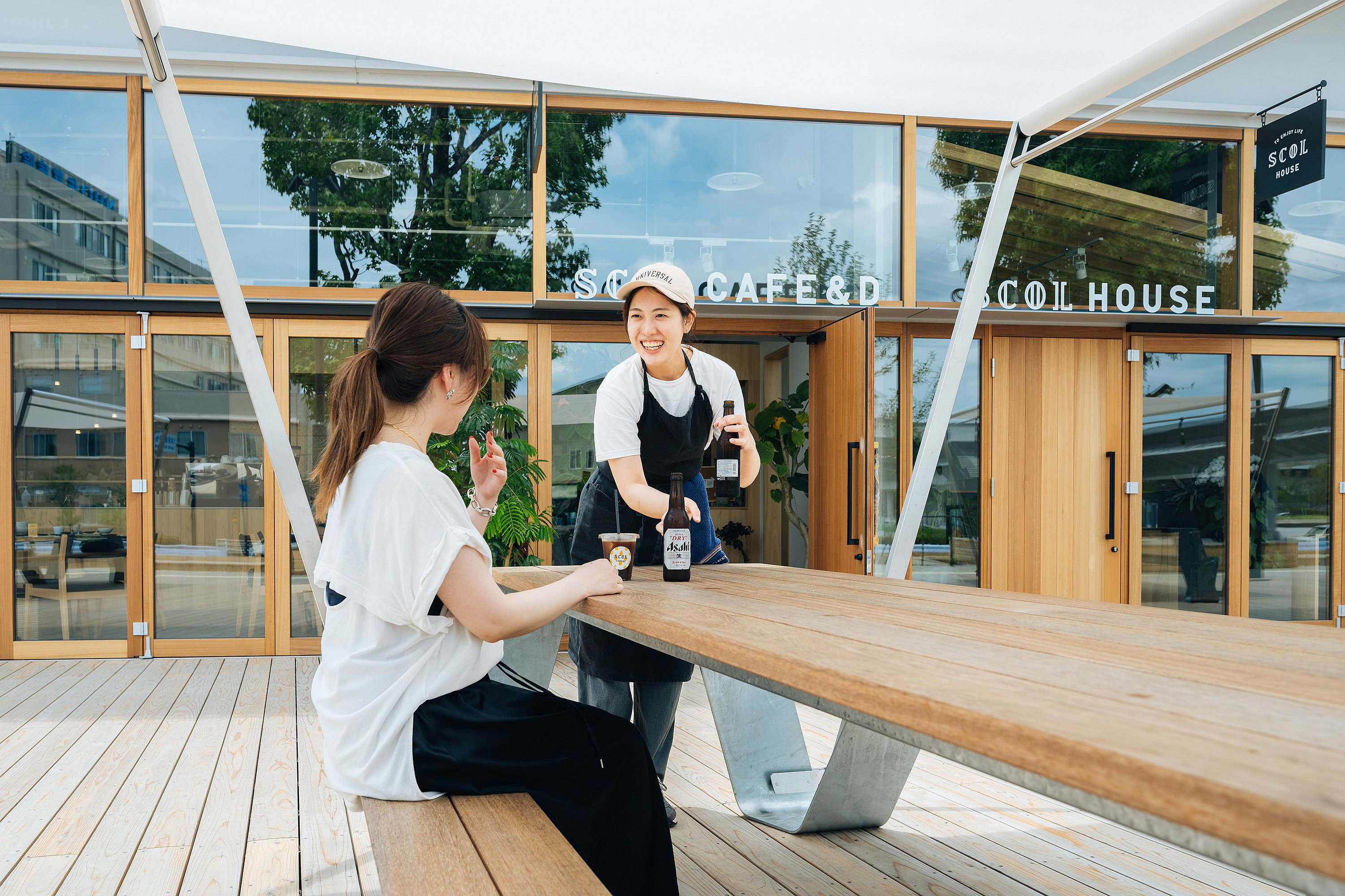 Staff member serving bottled drinks to a seated woman at a long wooden hopper picnic table in front of the SCOL HOUSE storefront with large glass doors and windows.