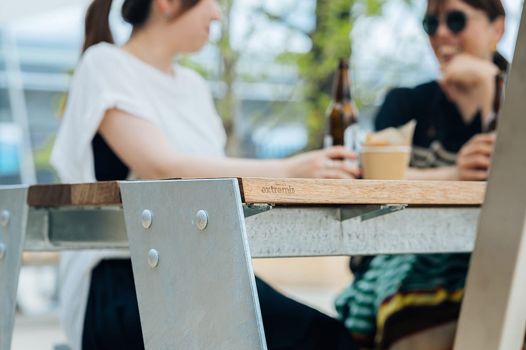 Close-up of a galvanized steel table frame and wooden tabletop edge with the Extremis logo, with two women talking over drinks in soft focus behind it.