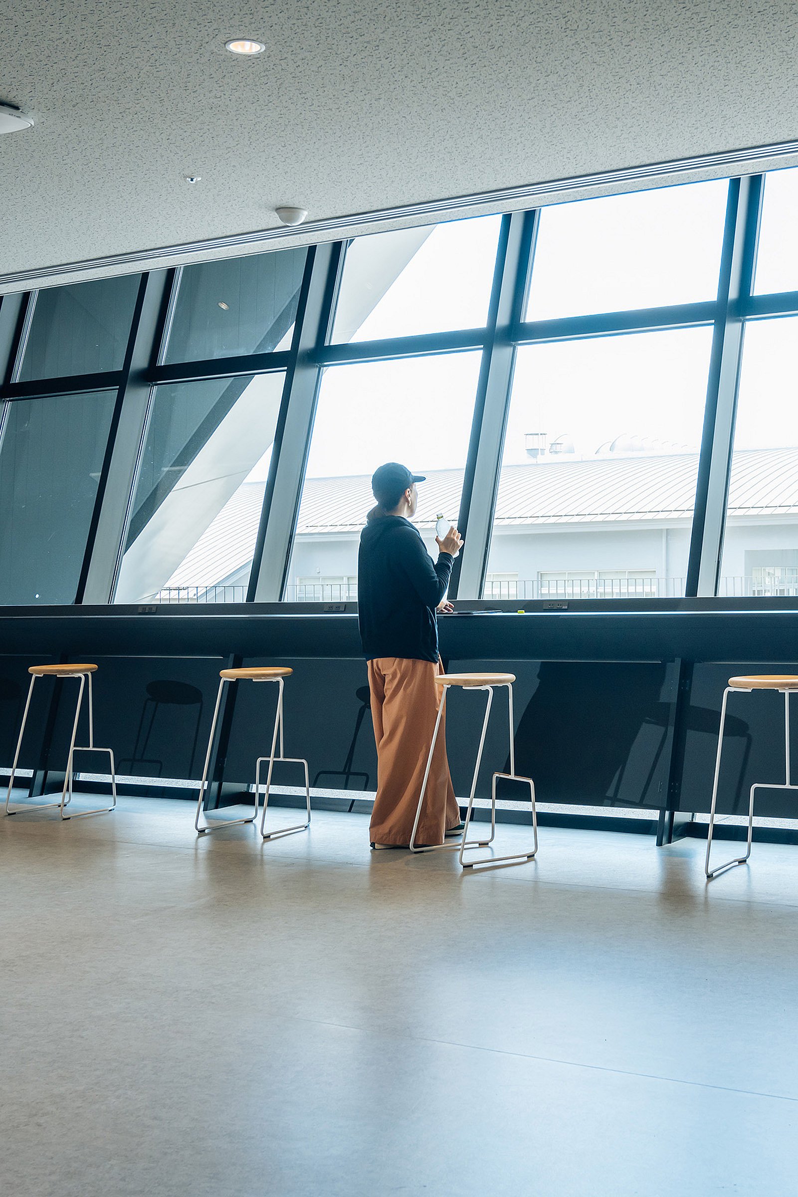 Person standing at a long counter and looking out through large slanted windows, with a row of tall tiki stools along the ledge.