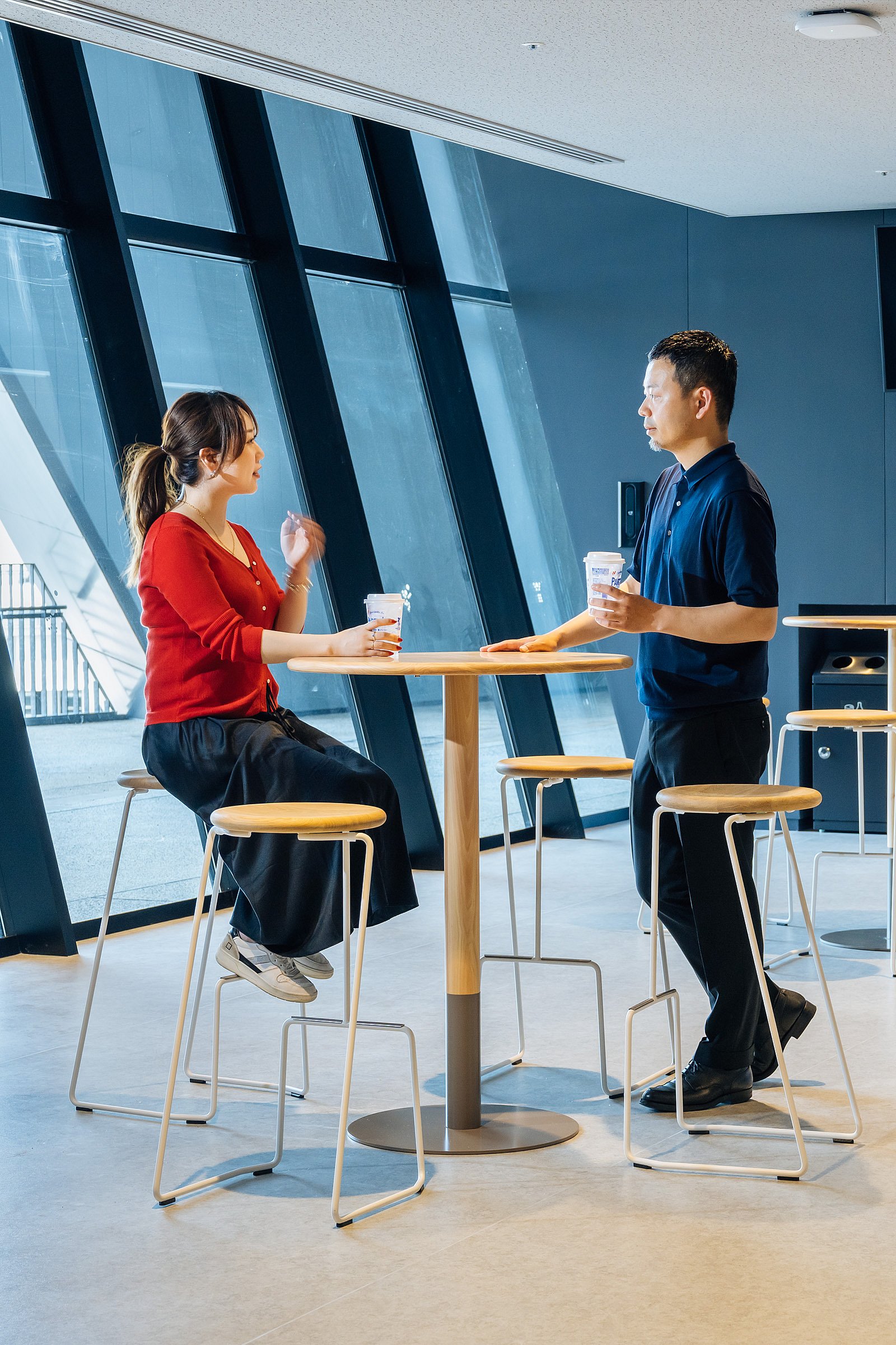 Woman and man talking at a tall round wooden tiki table with white wire-frame stools in front of large slanted floor-to-ceiling windows.